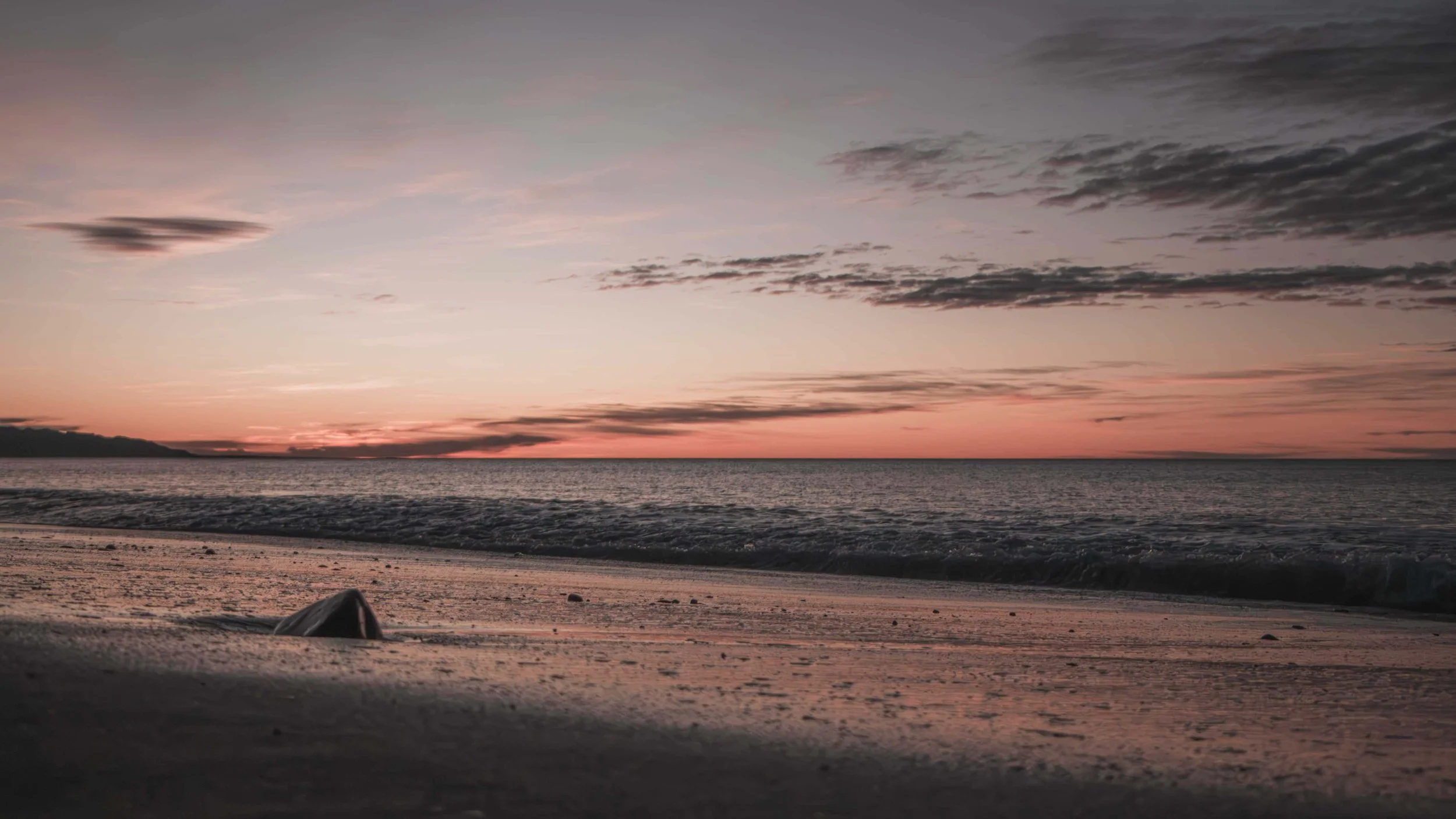 Sunset over the ocean with a sandy beach in the foreground, clouds in the sky, and a mountain in the distance.