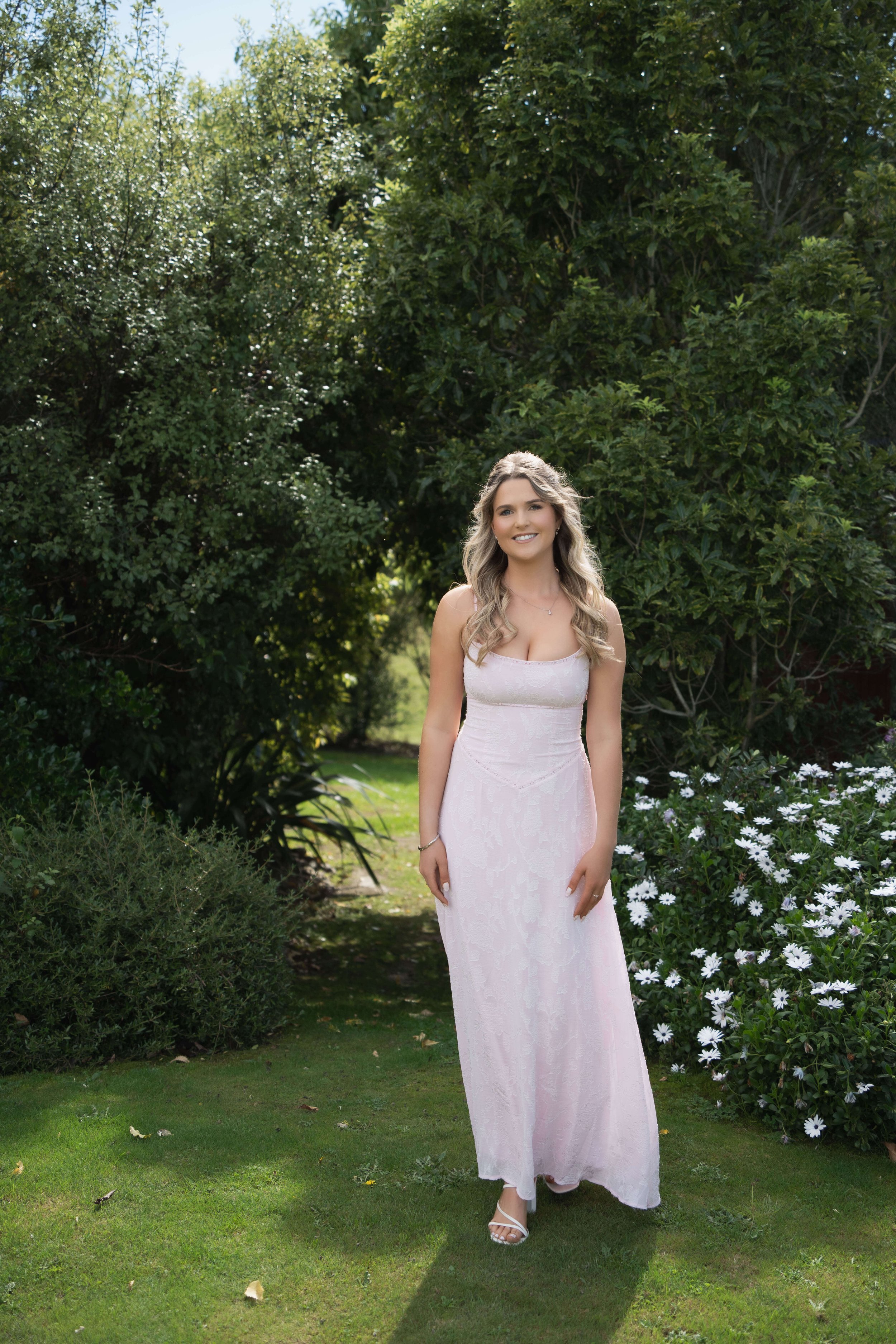A young woman in a light pink dress standing outdoors on a grassy path surrounded by green bushes and trees, smiling at the camera.