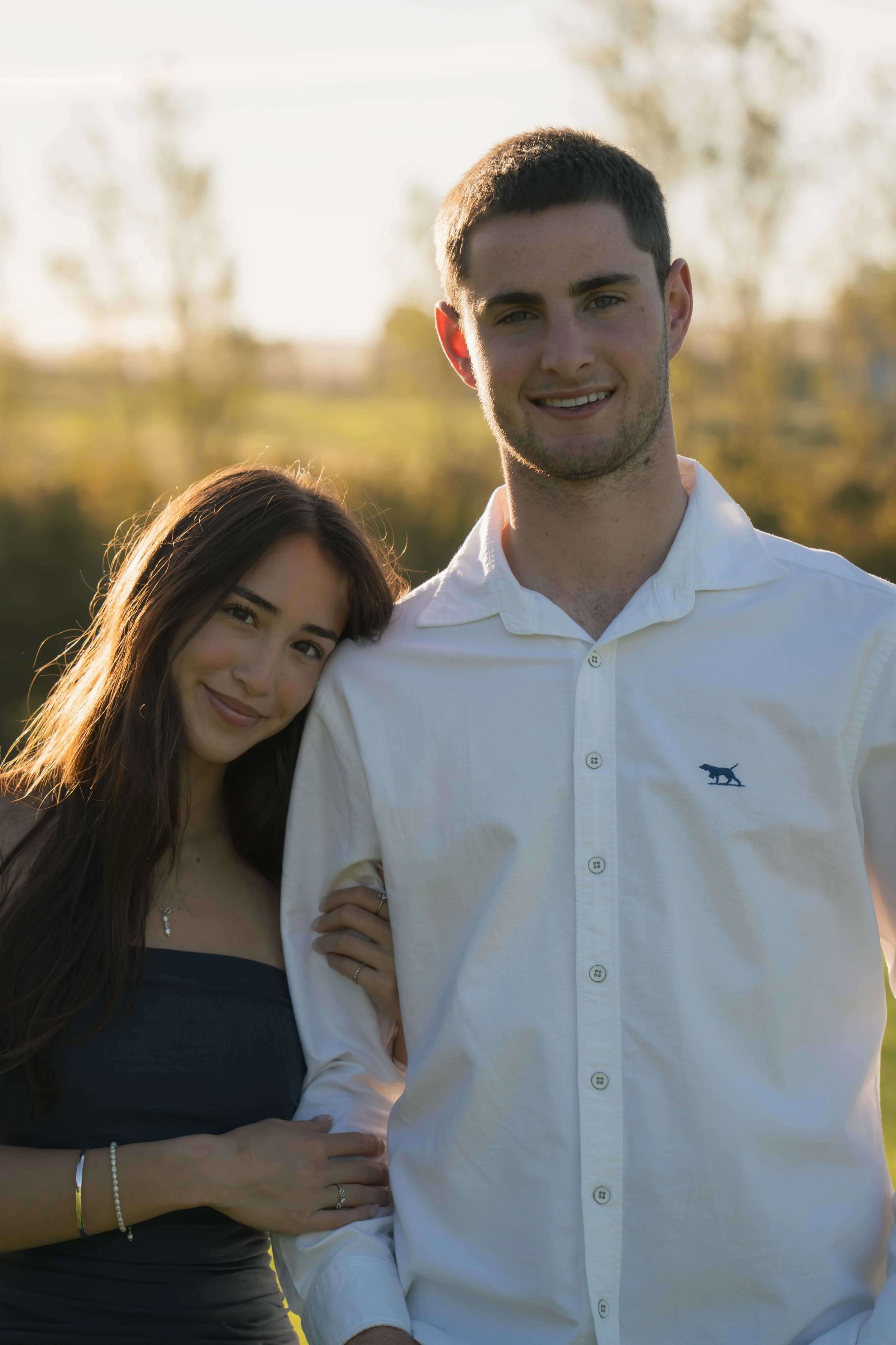 A young man and woman standing outdoors, smiling, with the woman leaning her head on the man's shoulder.