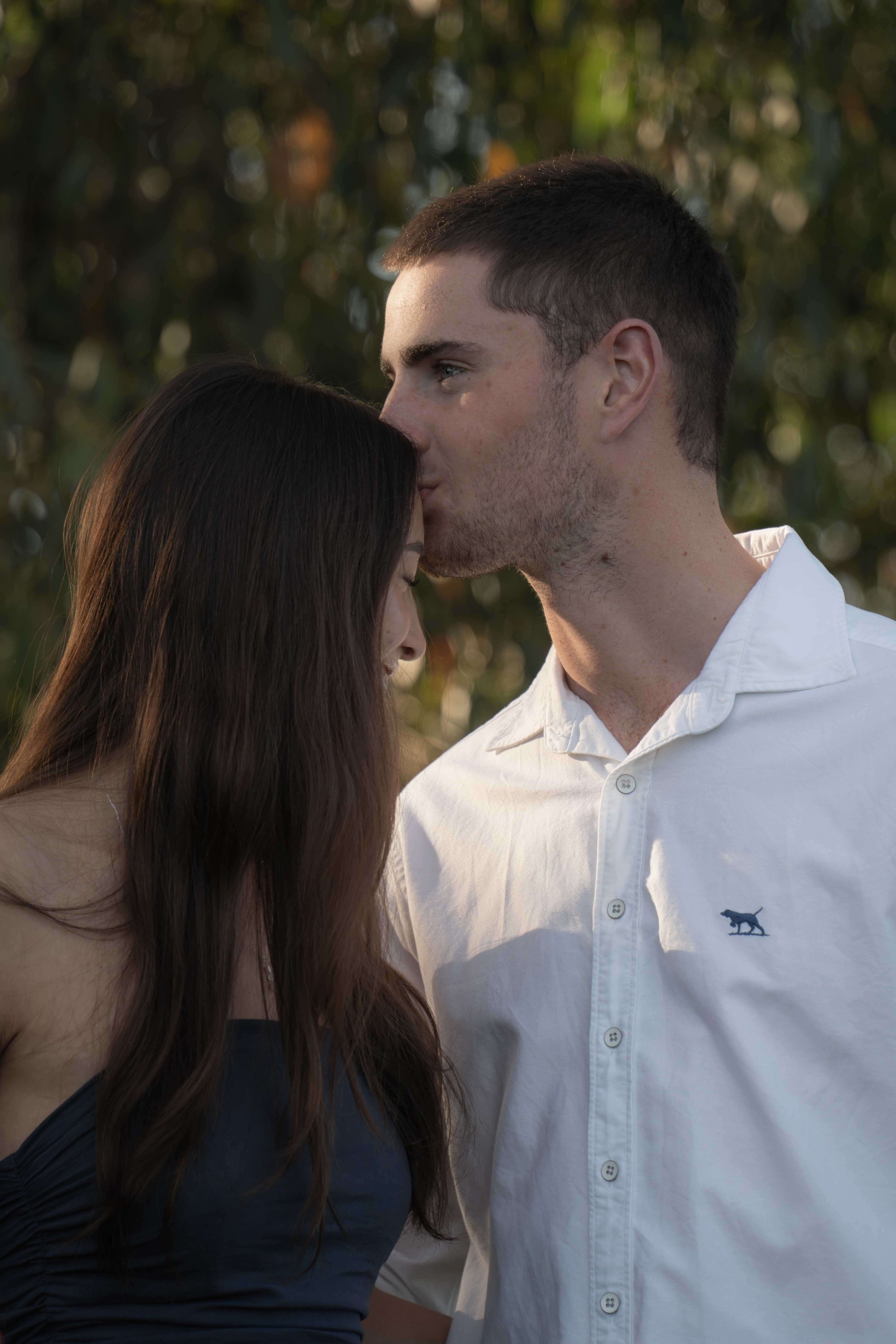 A man and woman share an intimate moment outdoors, with the man kissing the woman's forehead as she leans into the gesture.