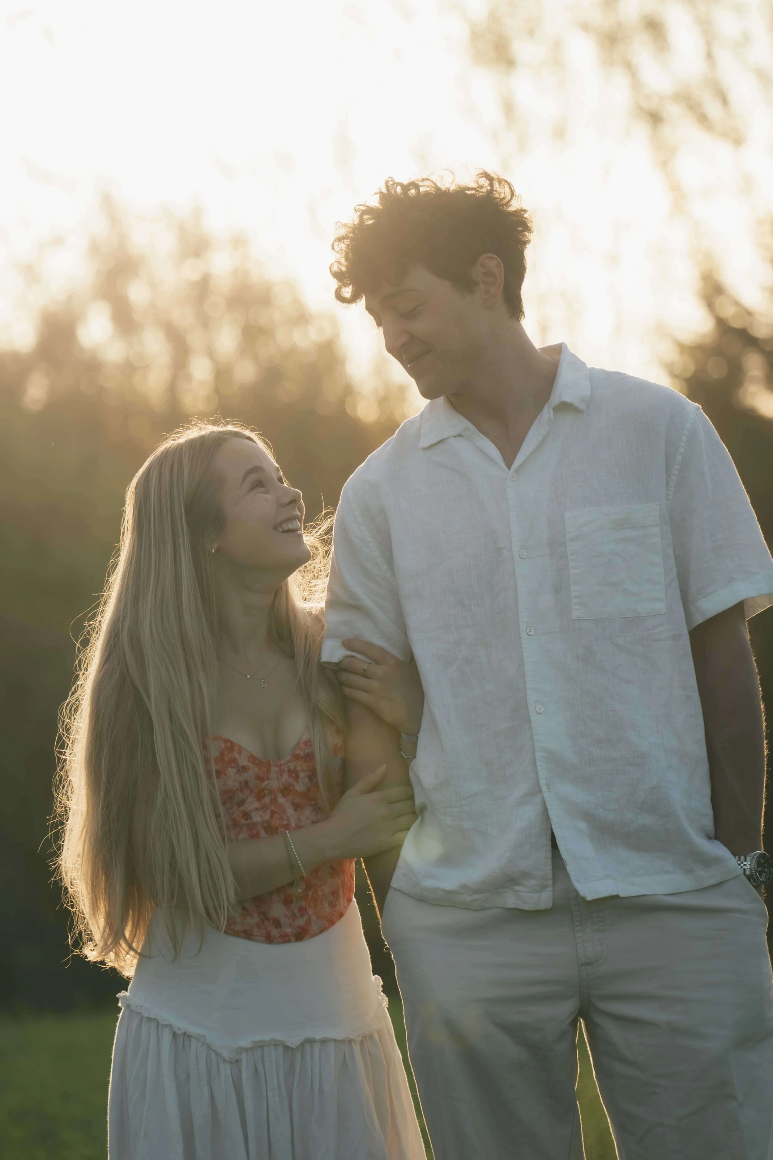 A young couple walking outdoors during sunset, smiling and looking at each other, with trees in the background.