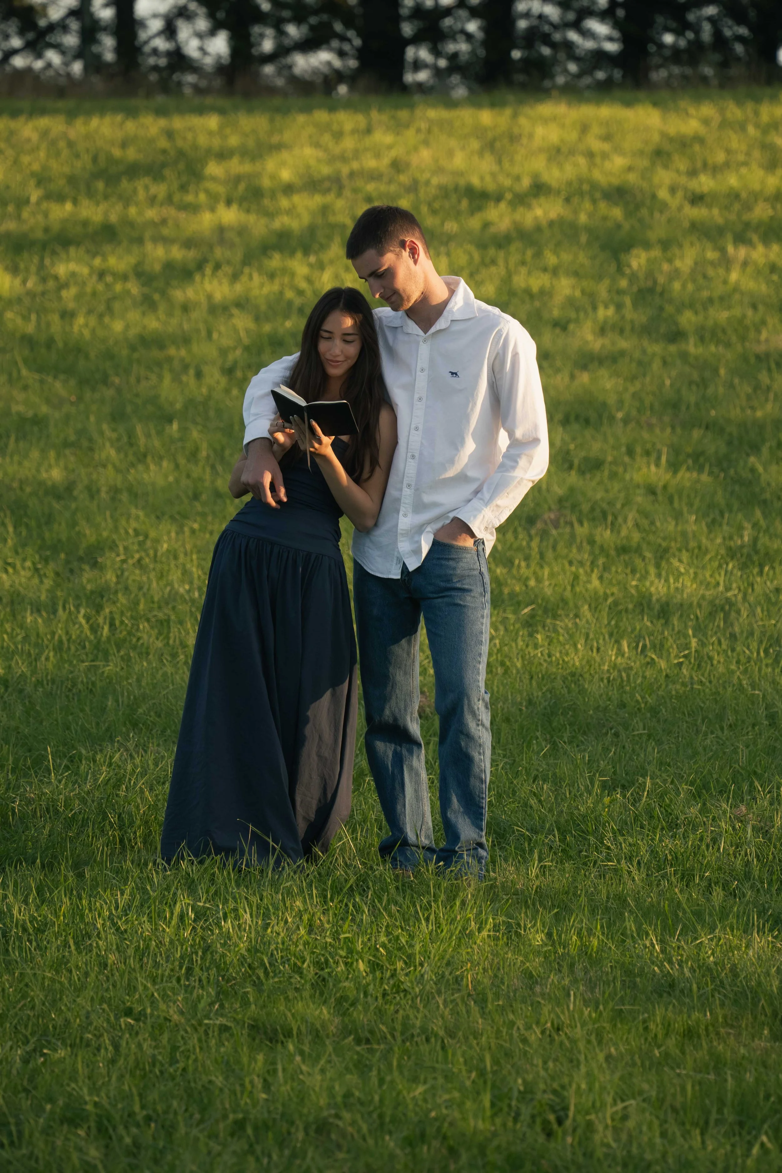 A young couple standing close together on a grassy field, looking at a small notebook or book together. The woman is wearing a dark dress, and the man is wearing a white shirt and jeans. They are outdoors during sunset or late afternoon.