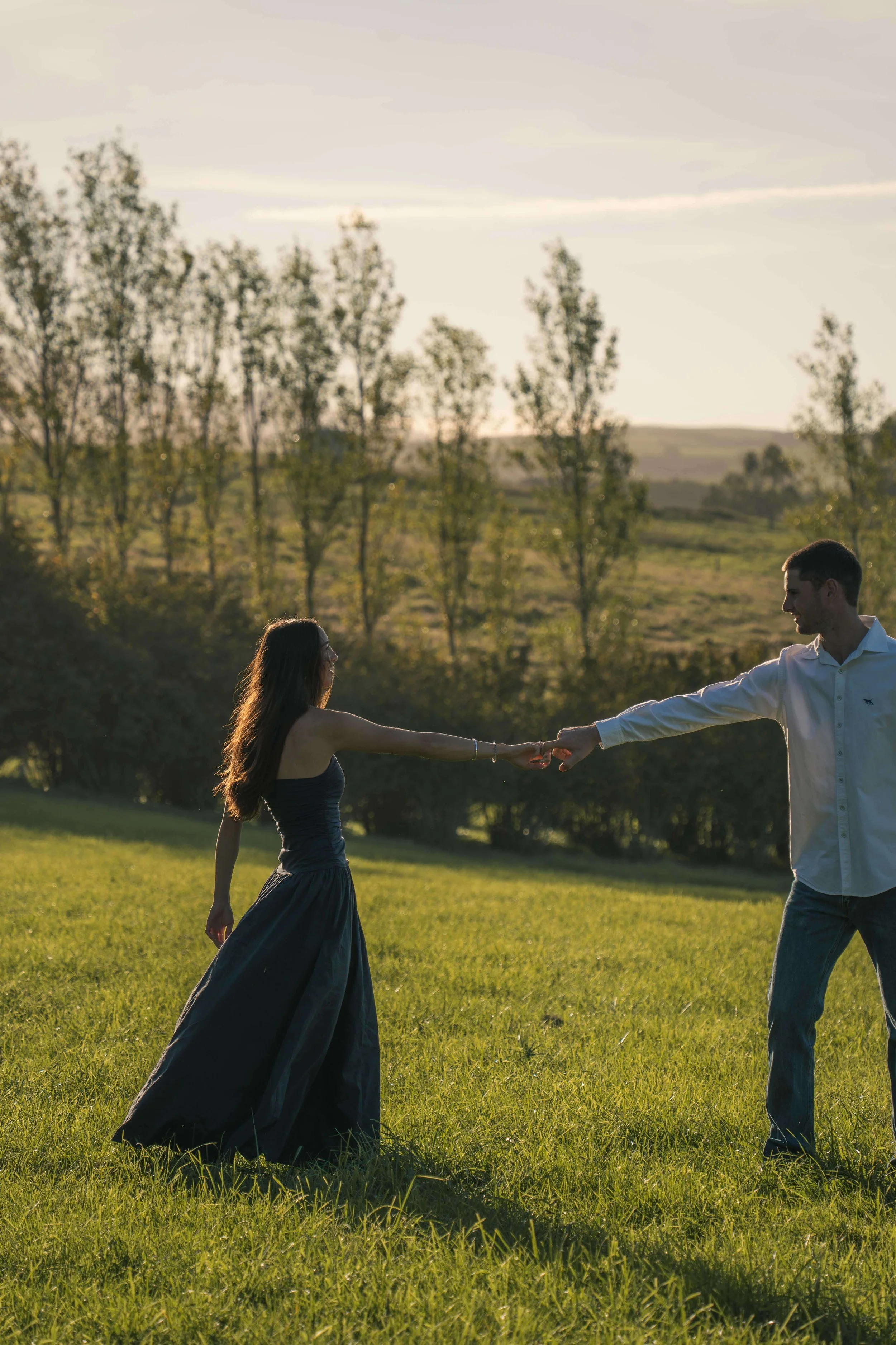 A couple dancing outdoors on a sunny day, holding hands in a grassy field with trees in the background.