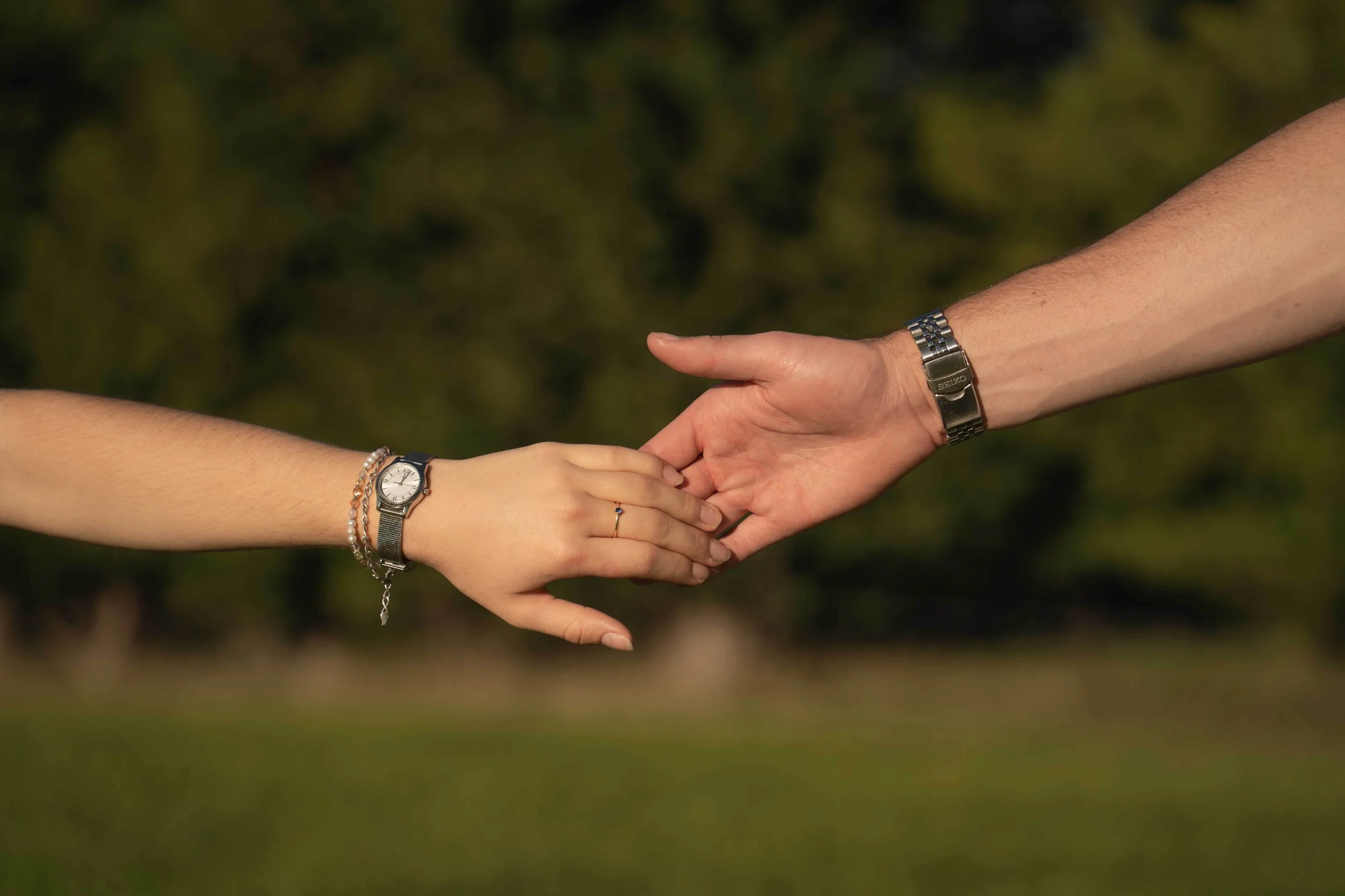 Close-up of two people holding hands, both wearing watches, outdoors with green foliage in the background.
