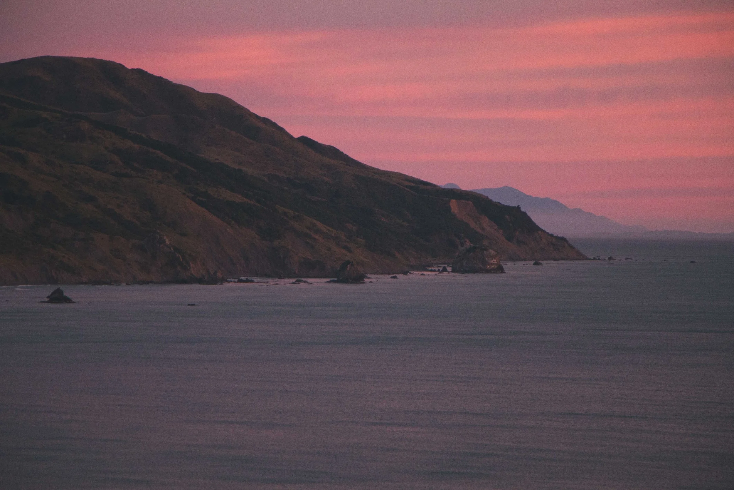 A scenic view of a coastline at dusk with a mountain range on the left and calm ocean water in the foreground, and pink clouds in the sky.