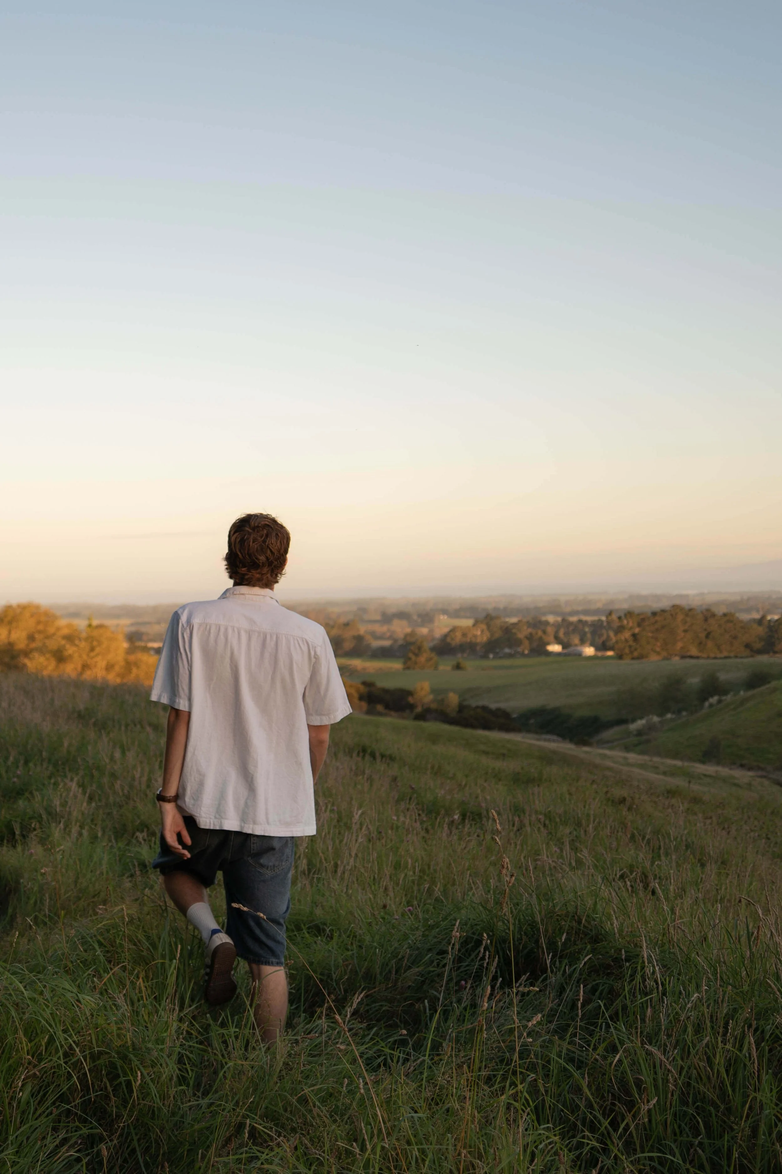 Person with curly hair wearing a white shirt, shorts, and sneakers walking through a grassy field during sunset, overlooking a landscape of hills and trees.