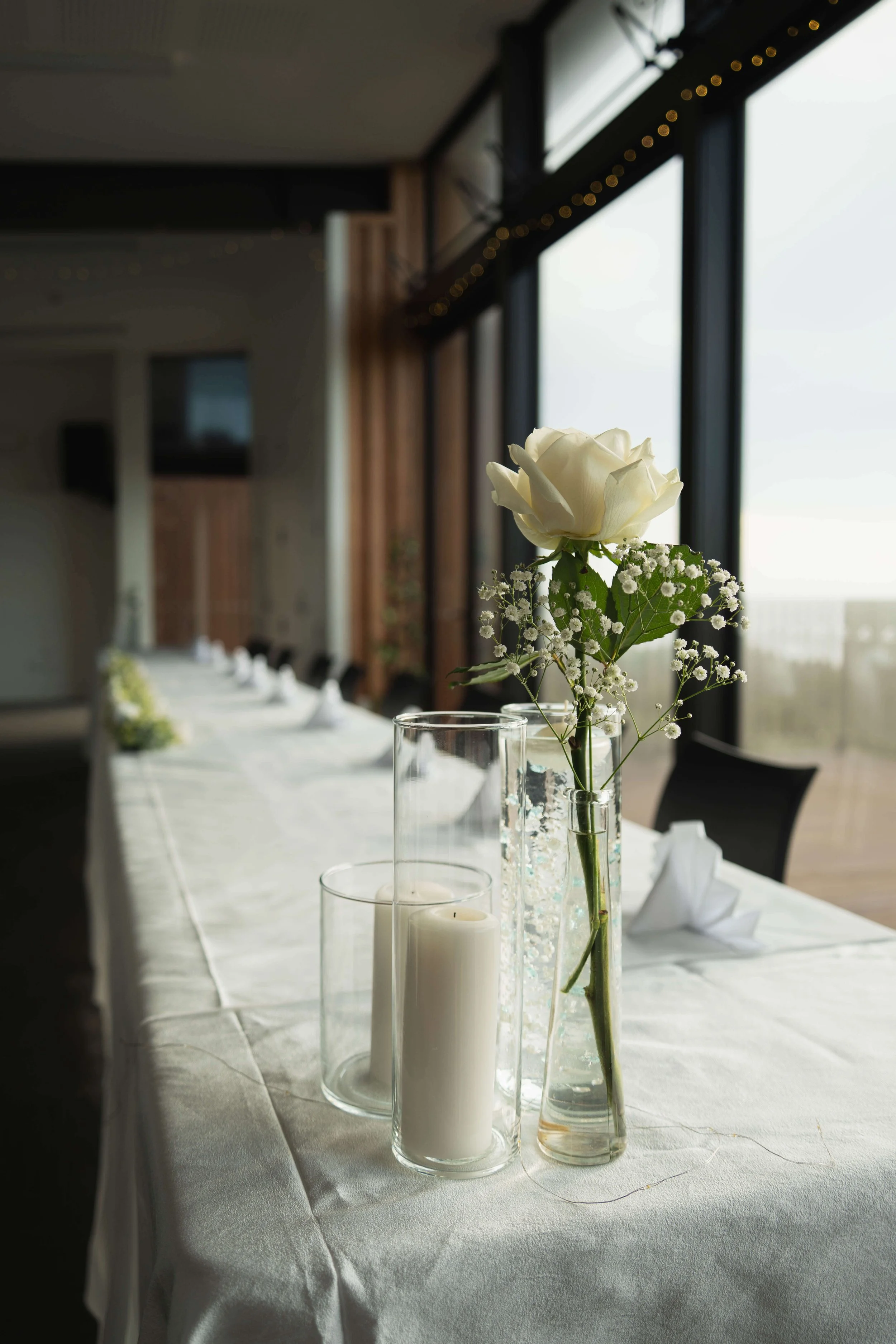 A close-up of a table centerpiece with white roses and baby's breath in a tall glass vase, accompanied by white candles in glass holders on a white tablecloth, with a large window and blurred indoor decor in the background.