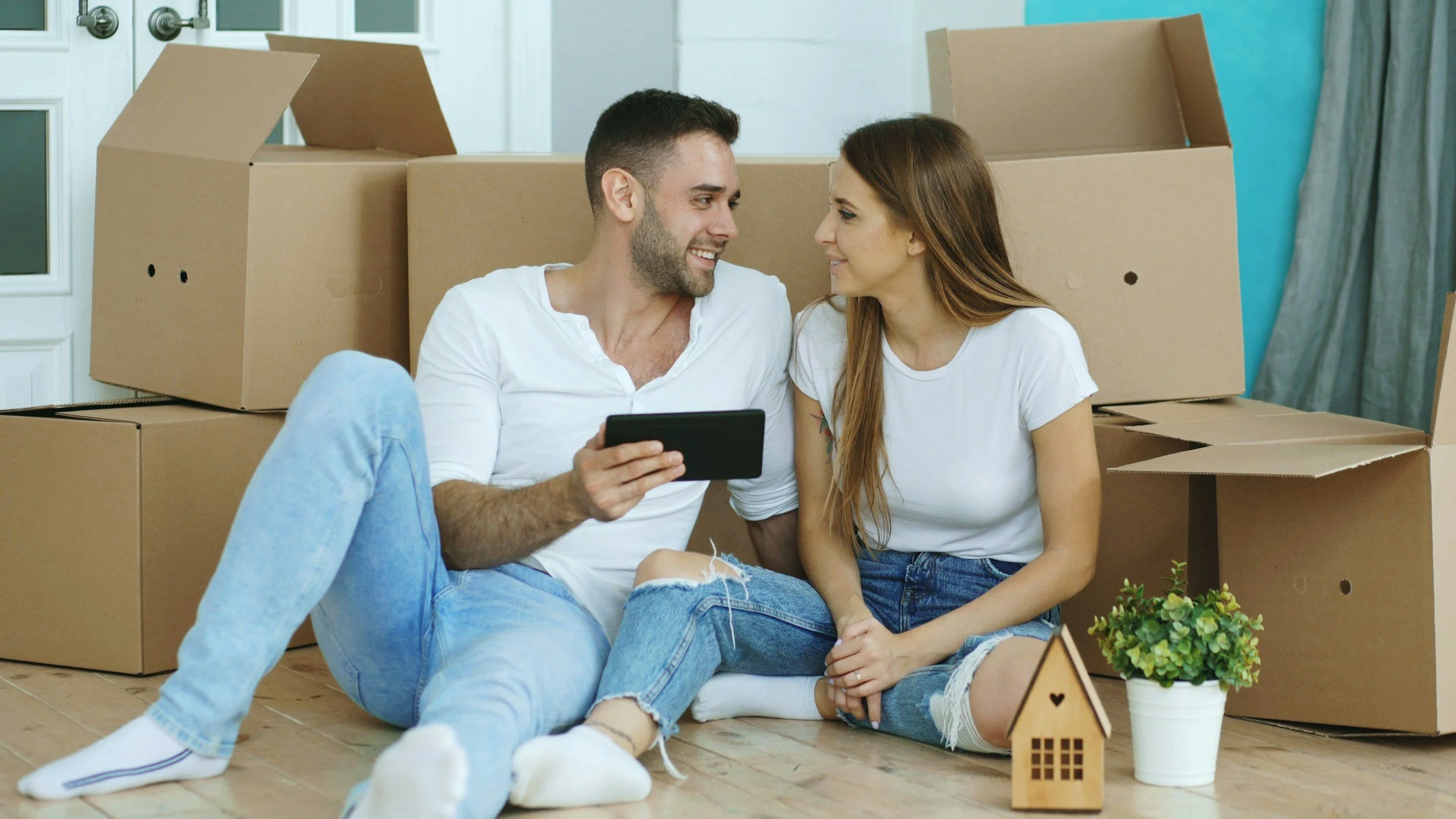 A young couple sitting on the floor amid moving boxes, smiling at each other, with a small model house and a potted plant in front.