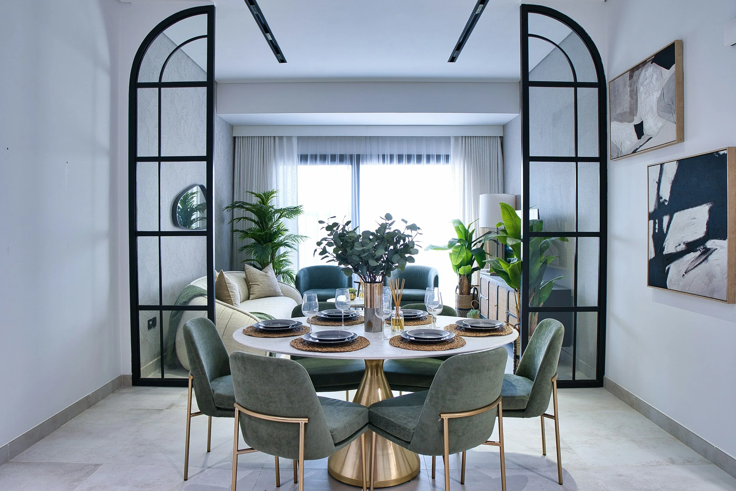 Modern dining room with a round marble table set with black dishes and glassware, surrounded by eight green upholstered chairs, and a bright living room with sofas, plants, and wall art in the background.