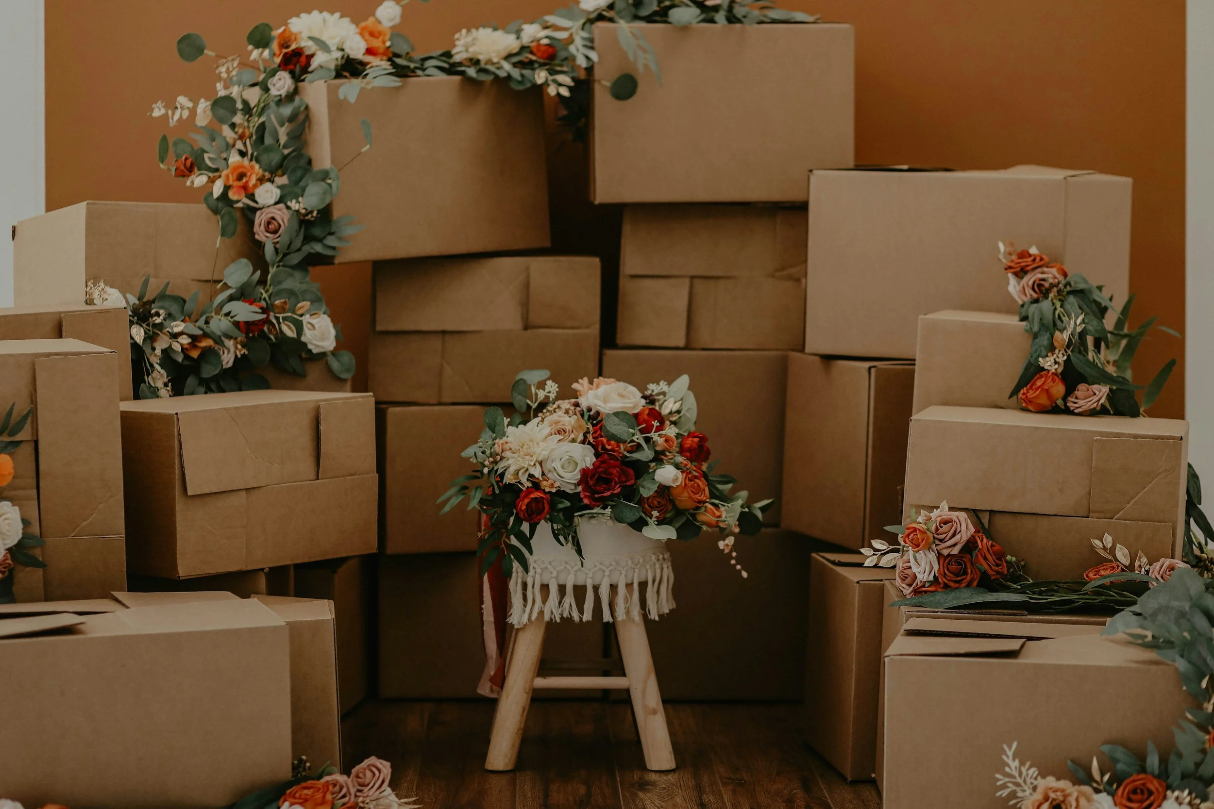 A display of cardboard boxes and floral arrangements, with a small wooden stool holding a bouquet of roses and greenery, set against a brown wall.
