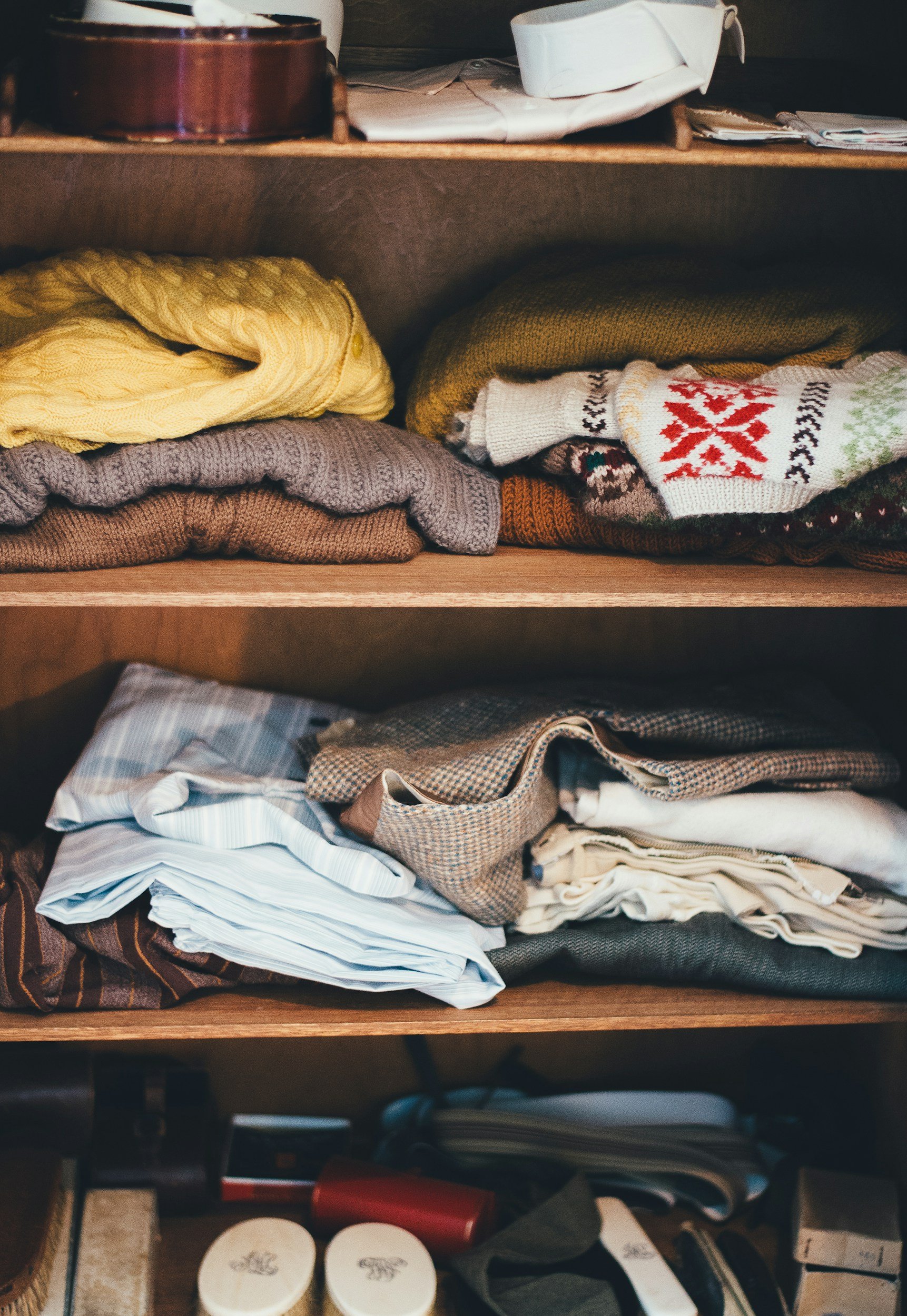 A wooden shelf with folded sweaters, shirts, and miscellaneous items, with some jars and personal items on the lower shelf.