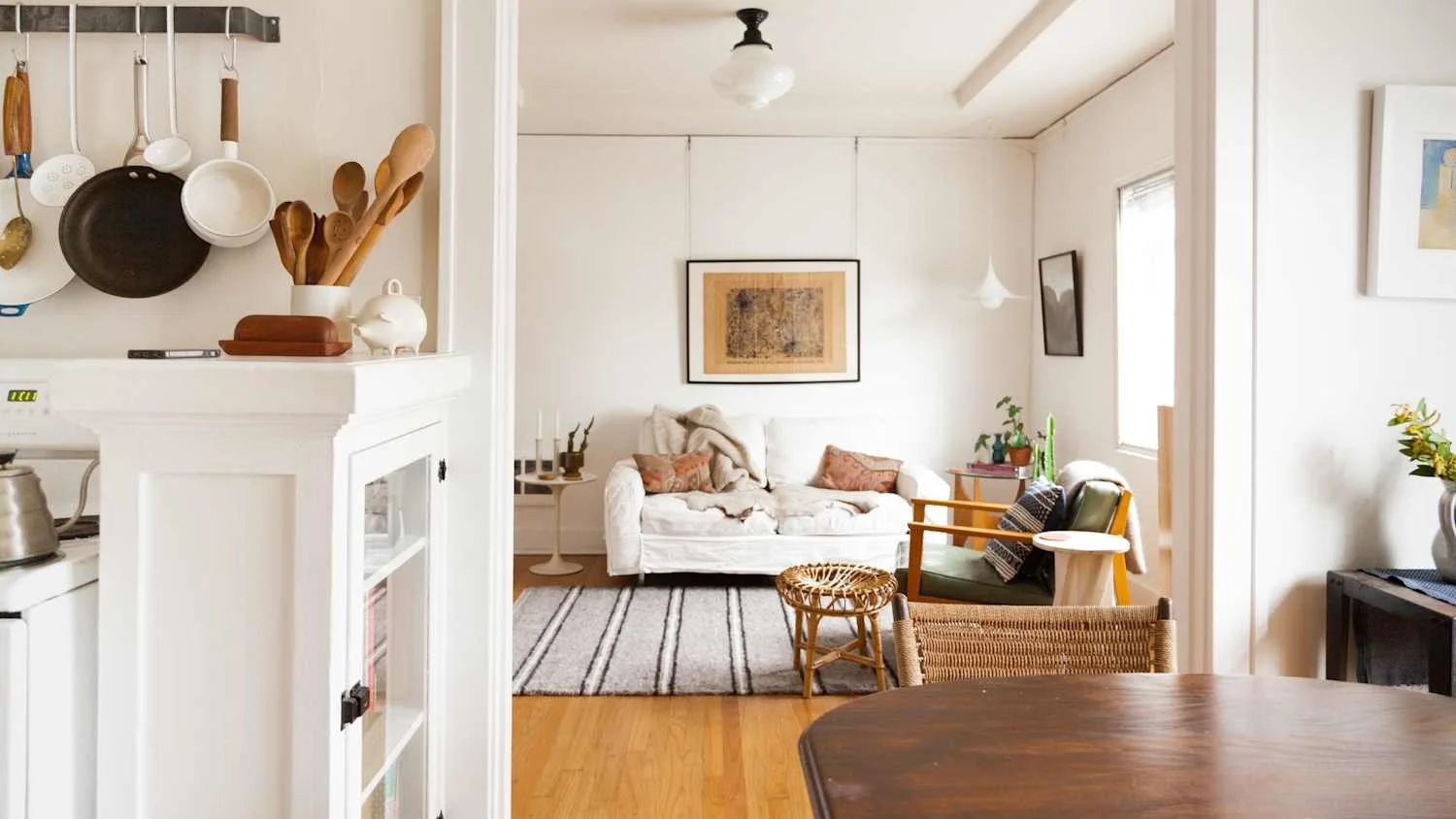 Living room with white sofa, green armchair, wooden side table, framed artwork, and striped rug, visible through an open doorway from a kitchen with hanging pots and utensils.