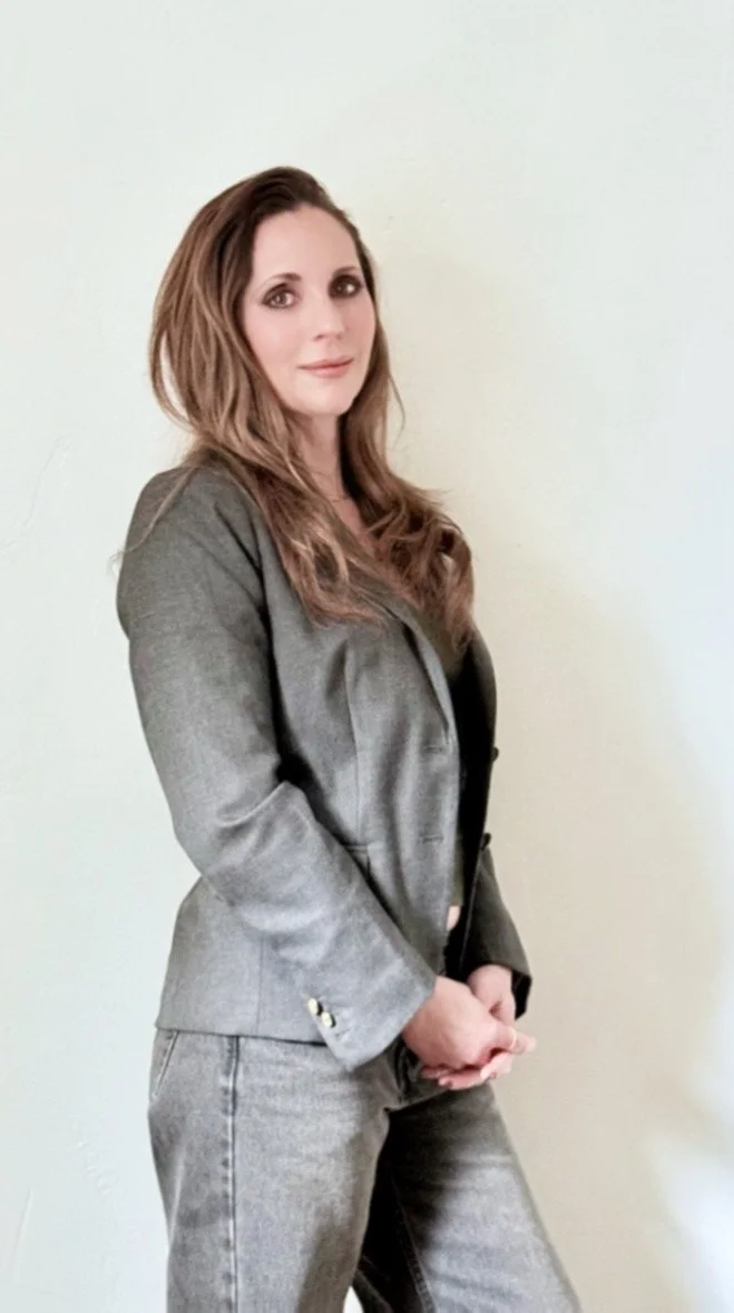 A woman with long, wavy light brown hair, wearing a gray blazer and matching jeans, standing against a plain light-colored wall.
