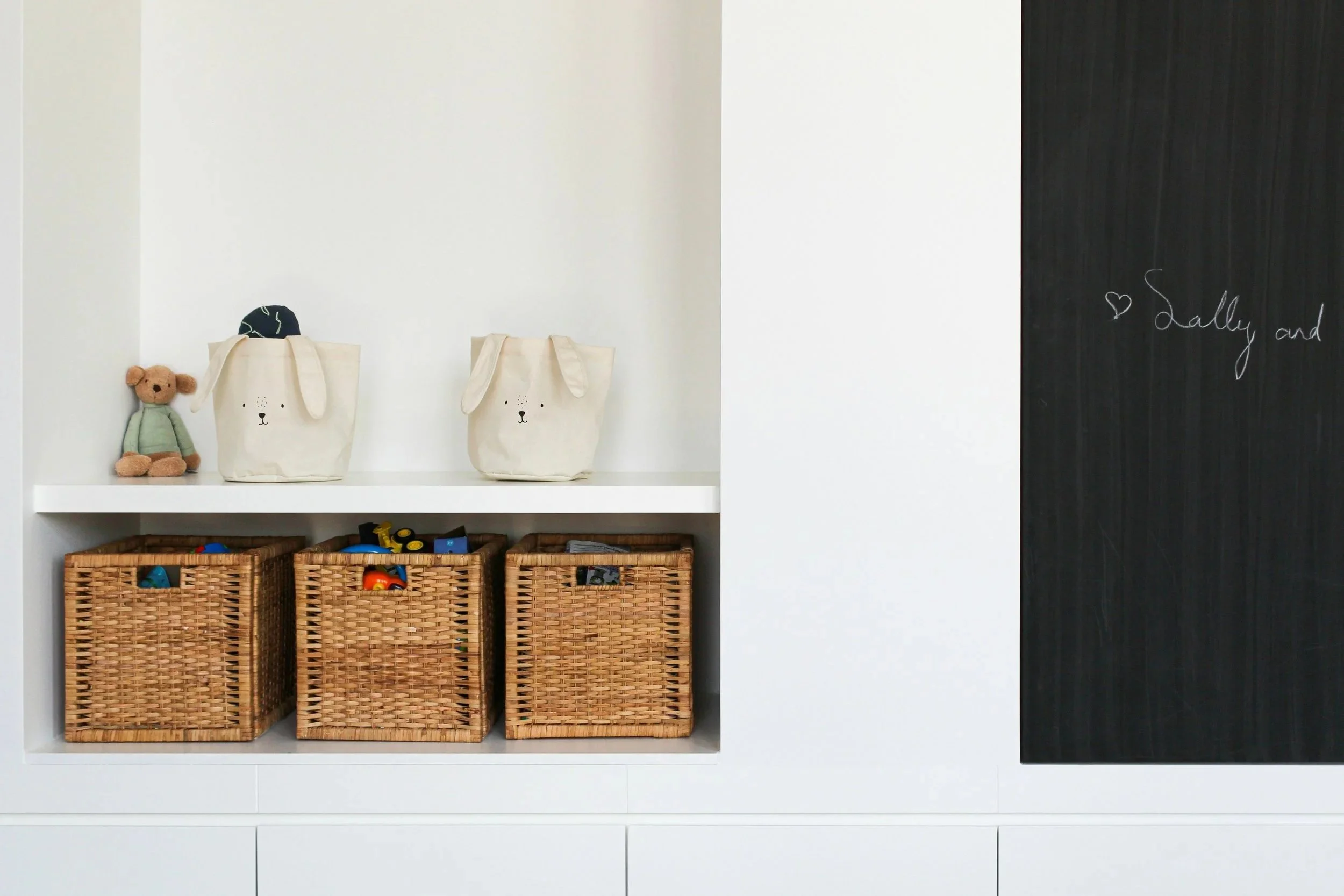 Children's toy shelf with stuffed bear, fabric bags with bunny faces, and woven baskets filled with toys, with a blackboard on the right side.
