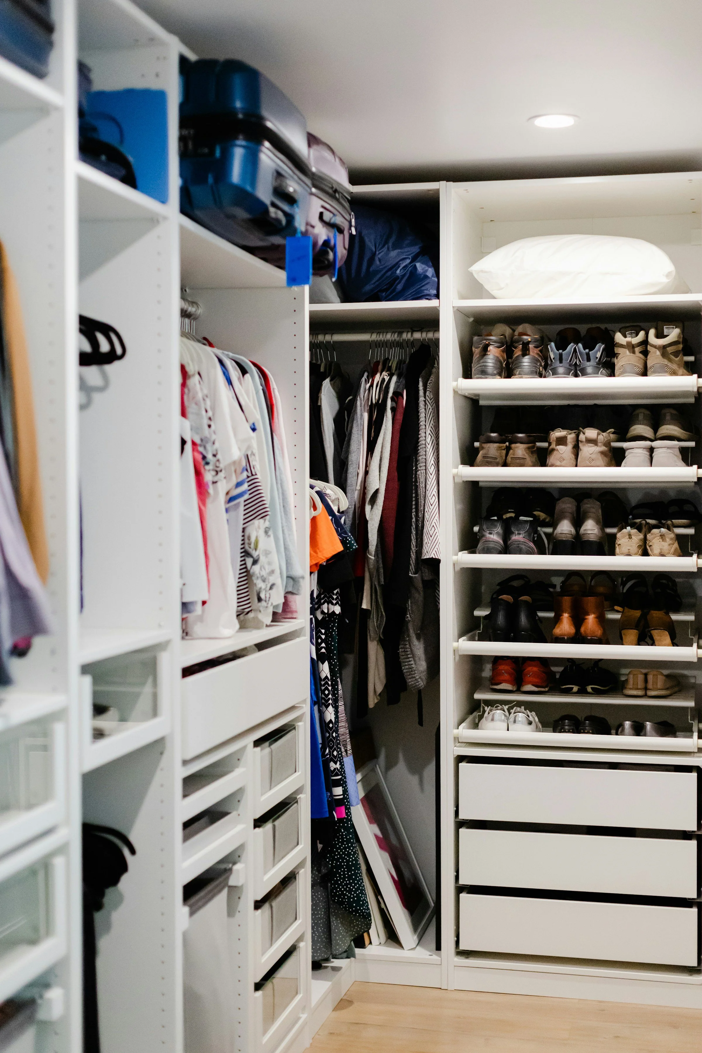 A walk-in closet with white shelving and drawers, containing shoes, clothing, luggage, and storage boxes.