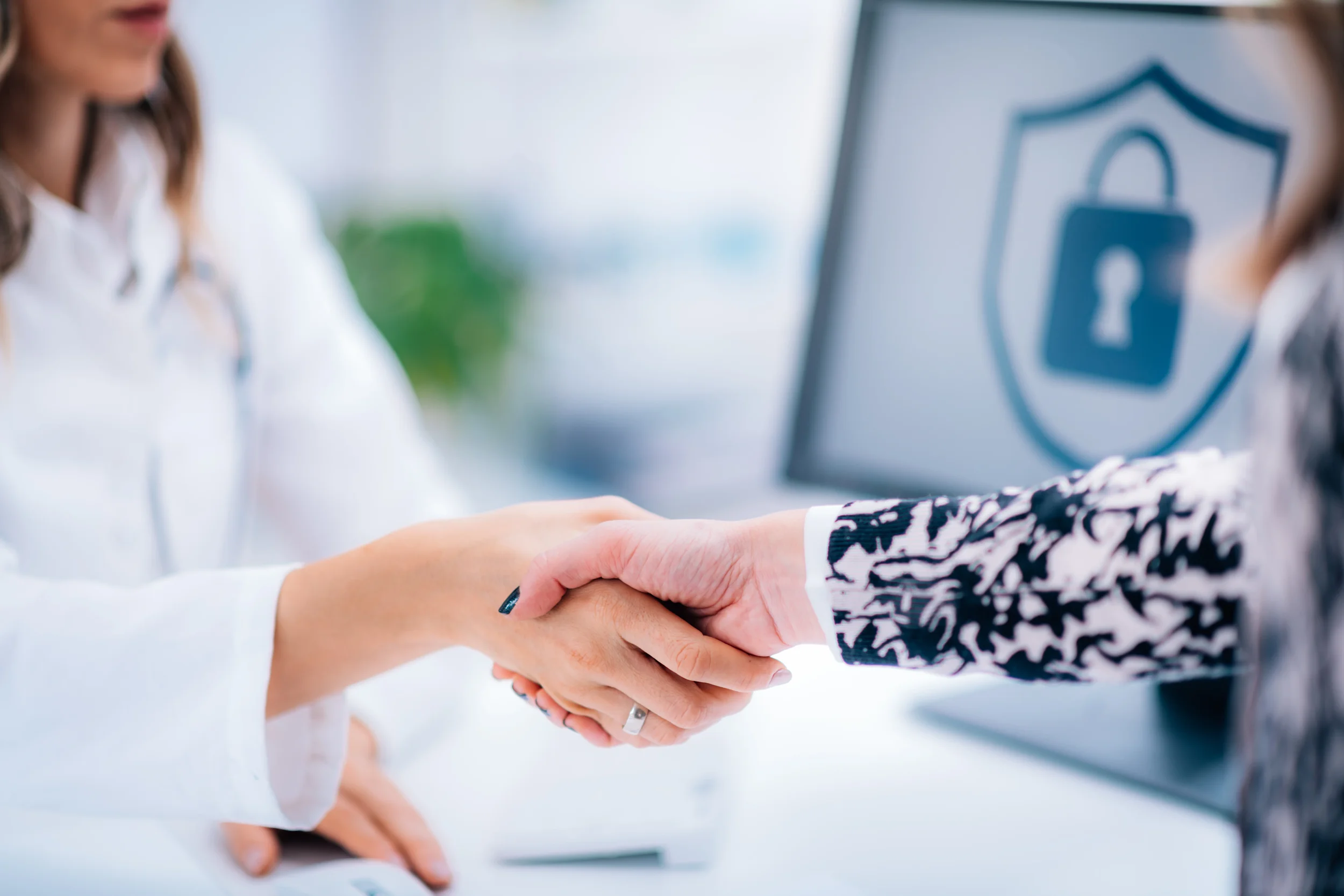 Two women shake hands in front of a computer with a security shield and lock icon, suggesting a business or cybersecurity agreement.