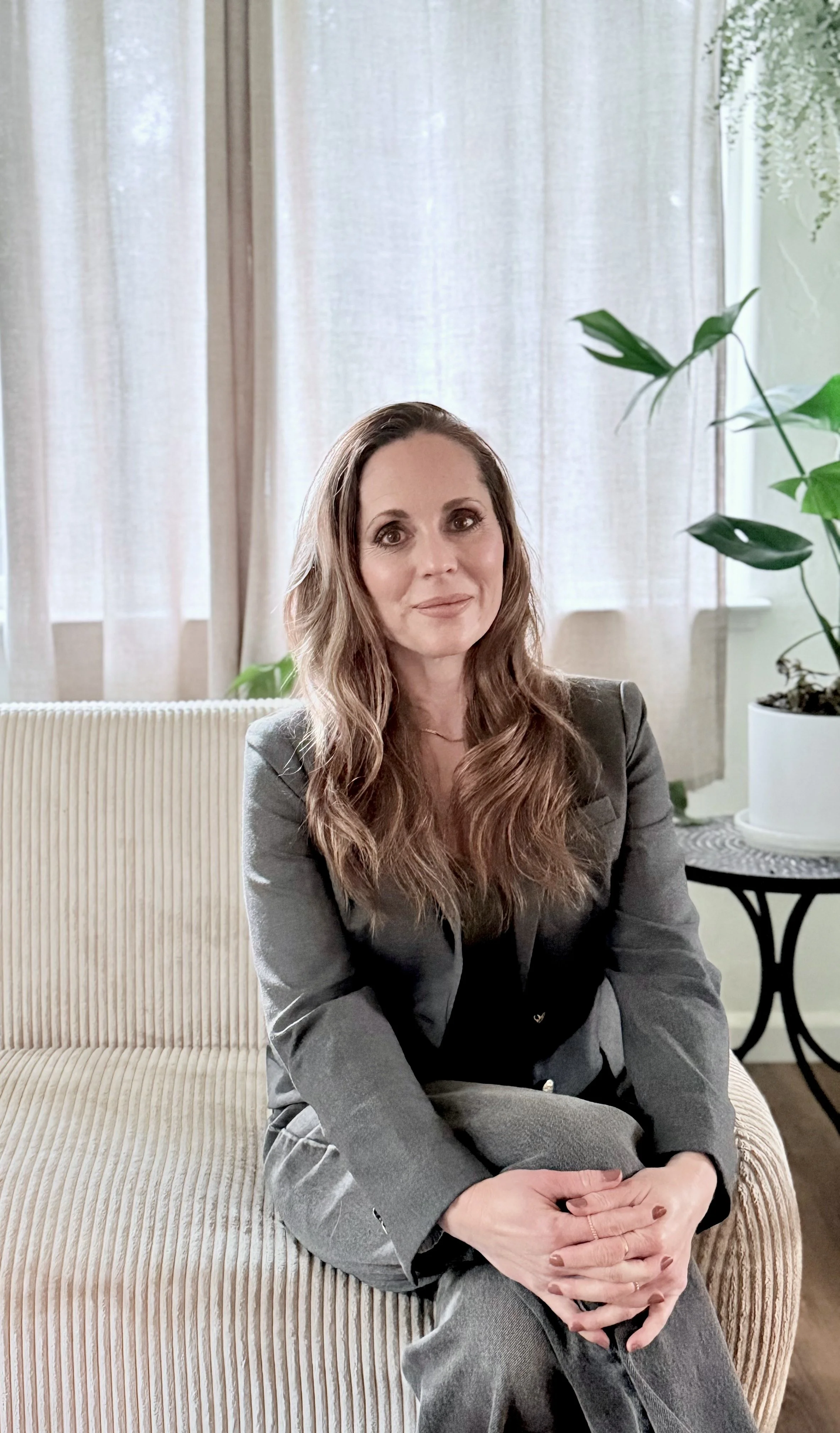 A woman with long wavy brown hair wearing a grey blazer and black top sitting on a beige corduroy couch in a well-lit room with white curtains and large green houseplants.