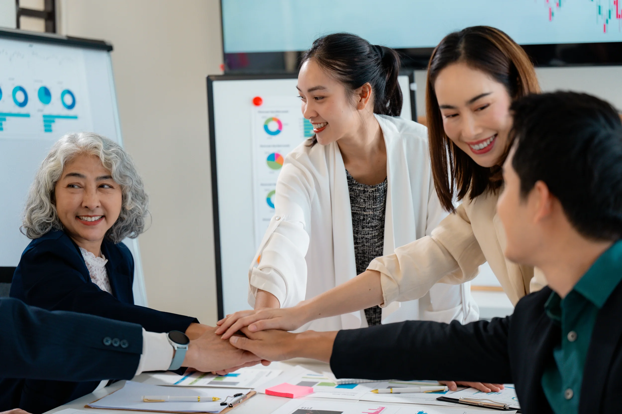 Five diverse professionals in a business meeting, smiling and placing their hands together on a table with charts and documents.