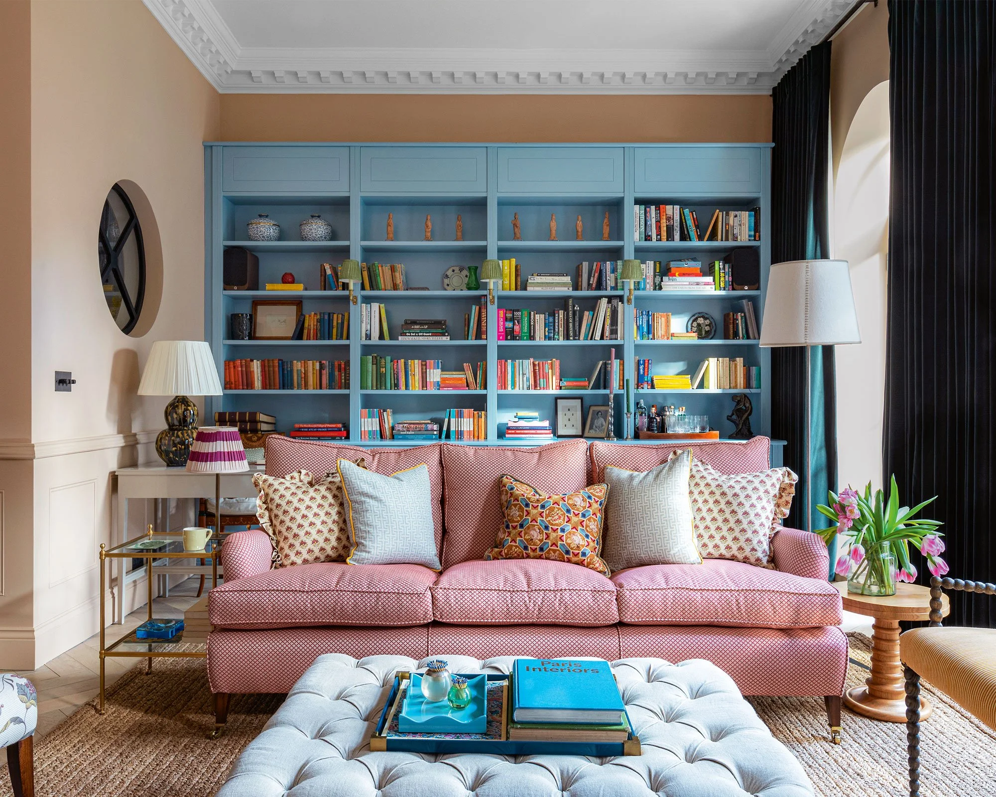 Living room with pink sofa, multiple colorful throw pillows, a cream tufted ottoman with books and decorative items, a side table with a lamp, a large blue built-in bookshelf filled with books and decorative objects, a window with dark curtains, and a vase of pink flowers on a wooden side table.