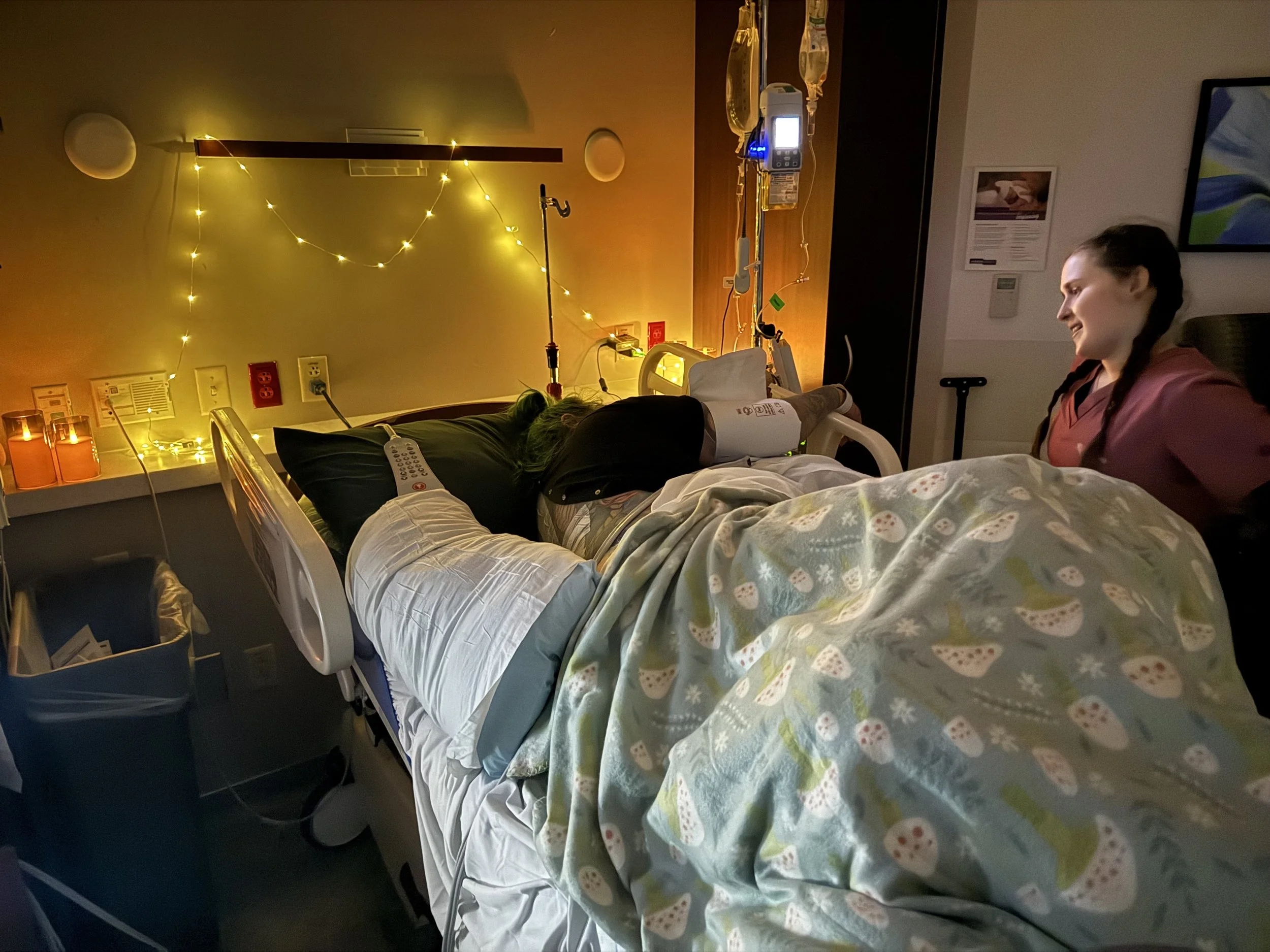 In a hospital room, a patient lying in bed connected to medical devices with a nurse sitting nearby, and a visitor smiling at the patient.