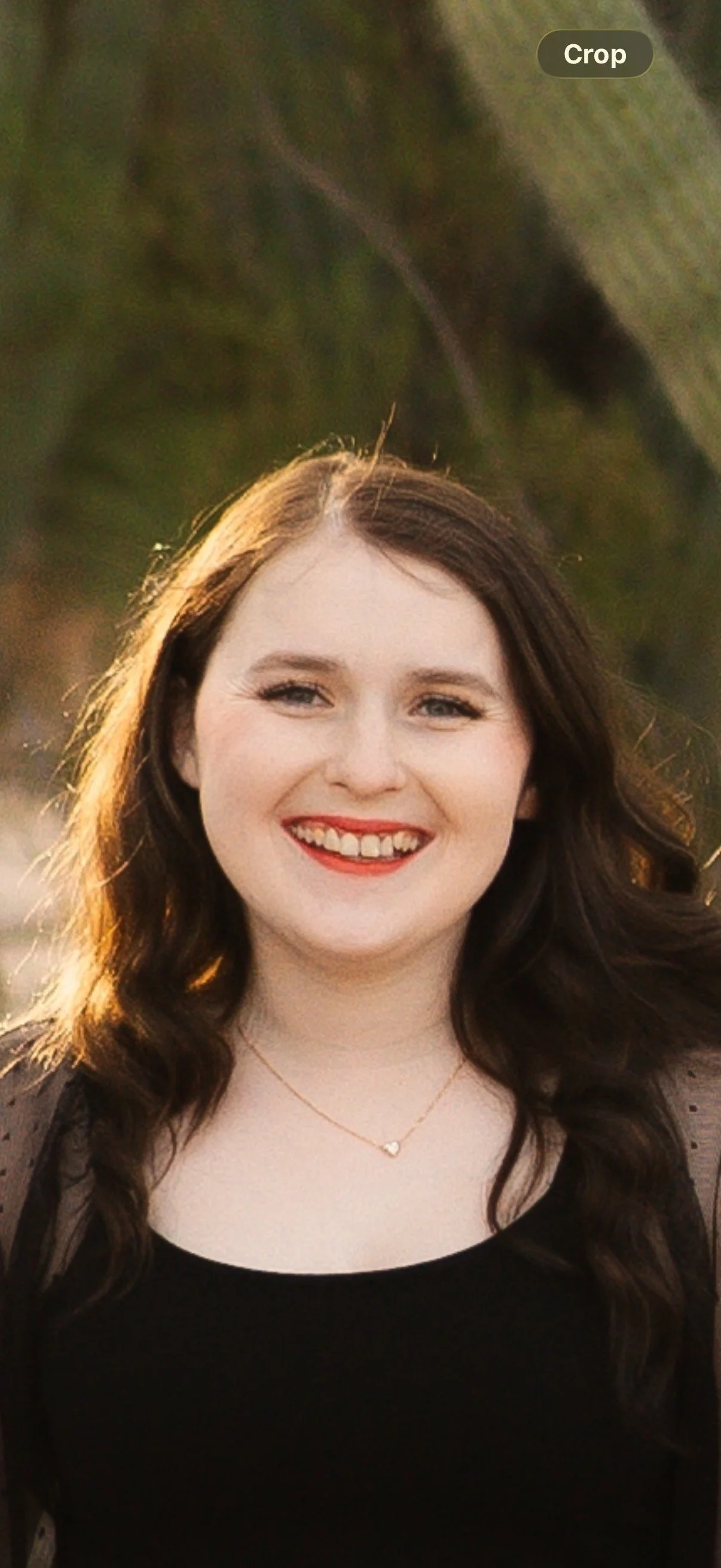 Portrait of a smiling young woman with long brown hair, wearing a black top and a delicate necklace, outdoors during golden hour with soft sunlight.