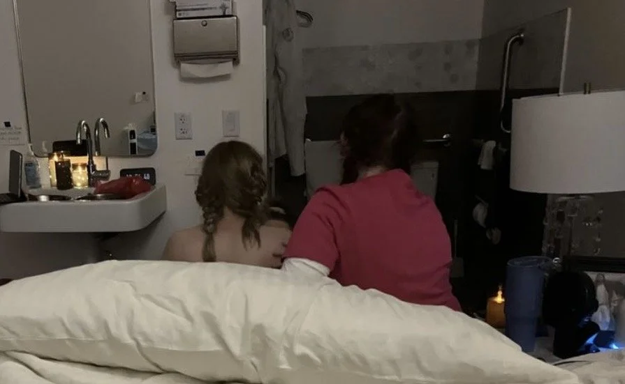 Two women sitting on a bed in a hotel room, facing away from the camera, with one wearing a red shirt and the other appearing topless.