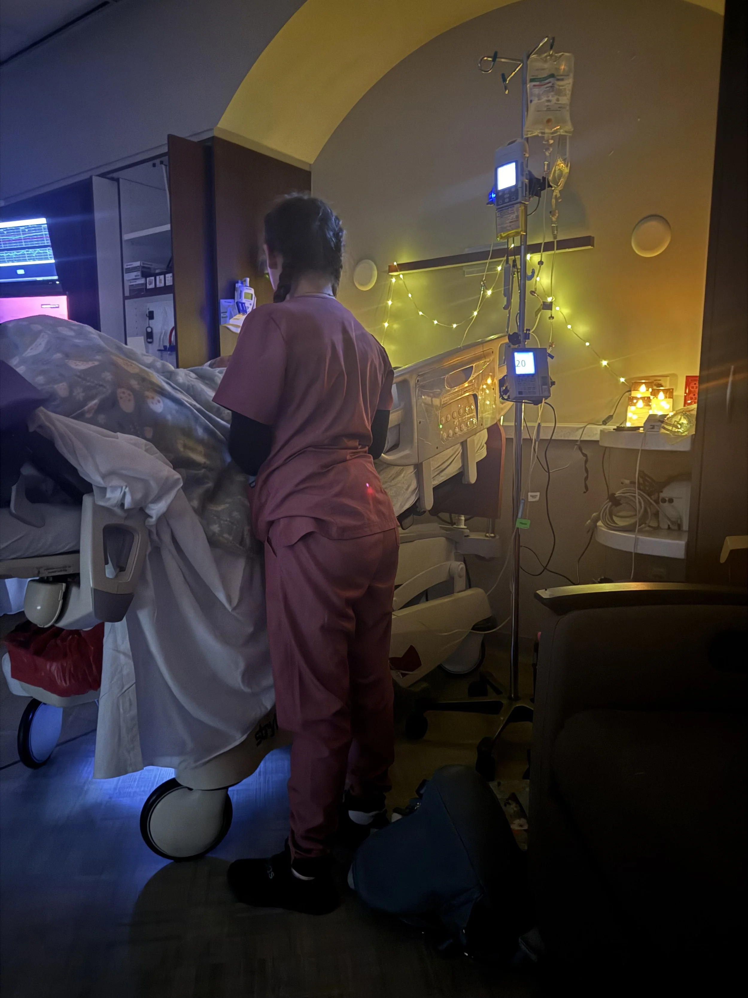A nurse in pink scrubs standing beside a patient bed in a hospital room, with medical equipment, IV drip, and monitors visible, illuminated by soft yellow and blue lighting.