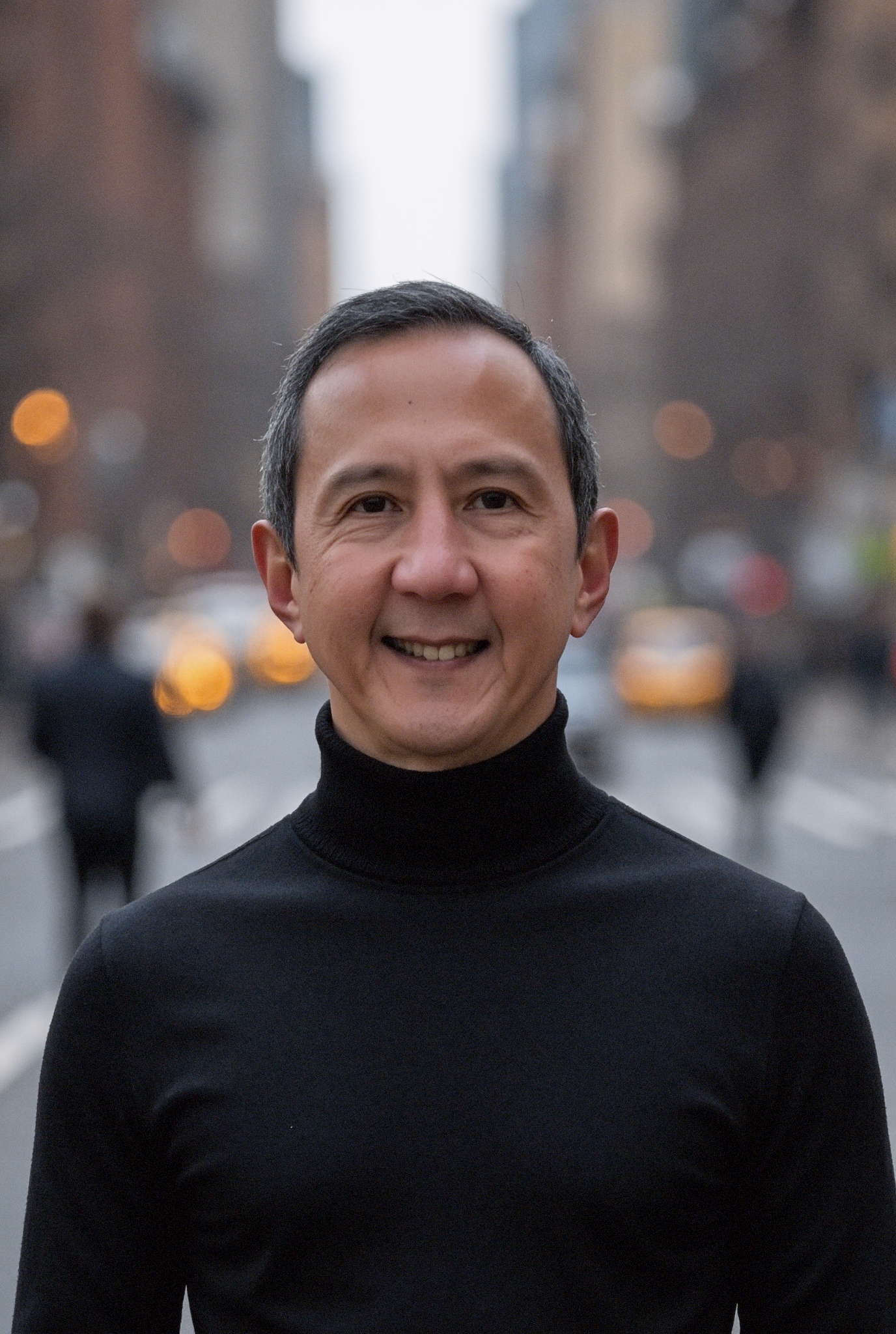 A smiling man in a black turtleneck sweater standing outdoors in an urban area with blurred buildings and city lights in the background.