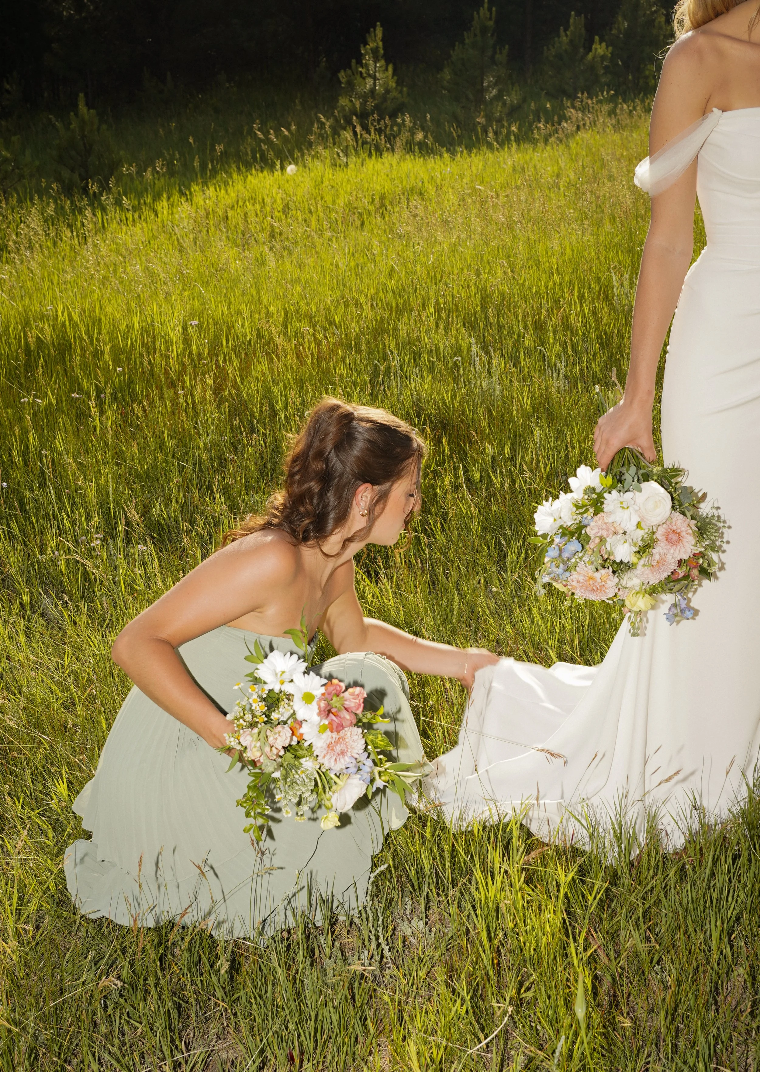 Candid bridesmaid adjusting brides dress in a grassy field holding her bouquet during a mountain wedding in Pine, Colorado.