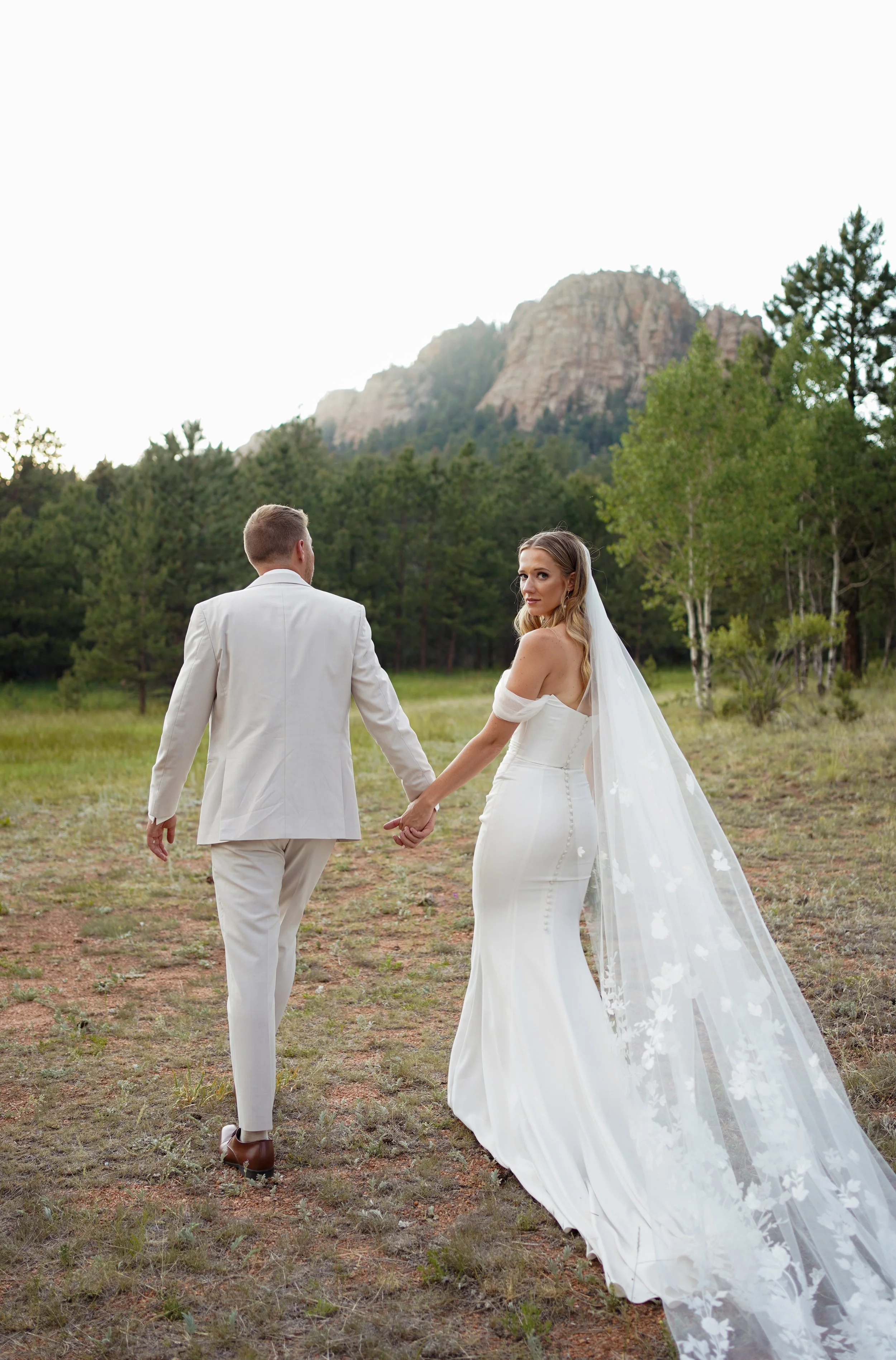 Bride and groom walking in a field with mountains and long veil in Pine, Colorado