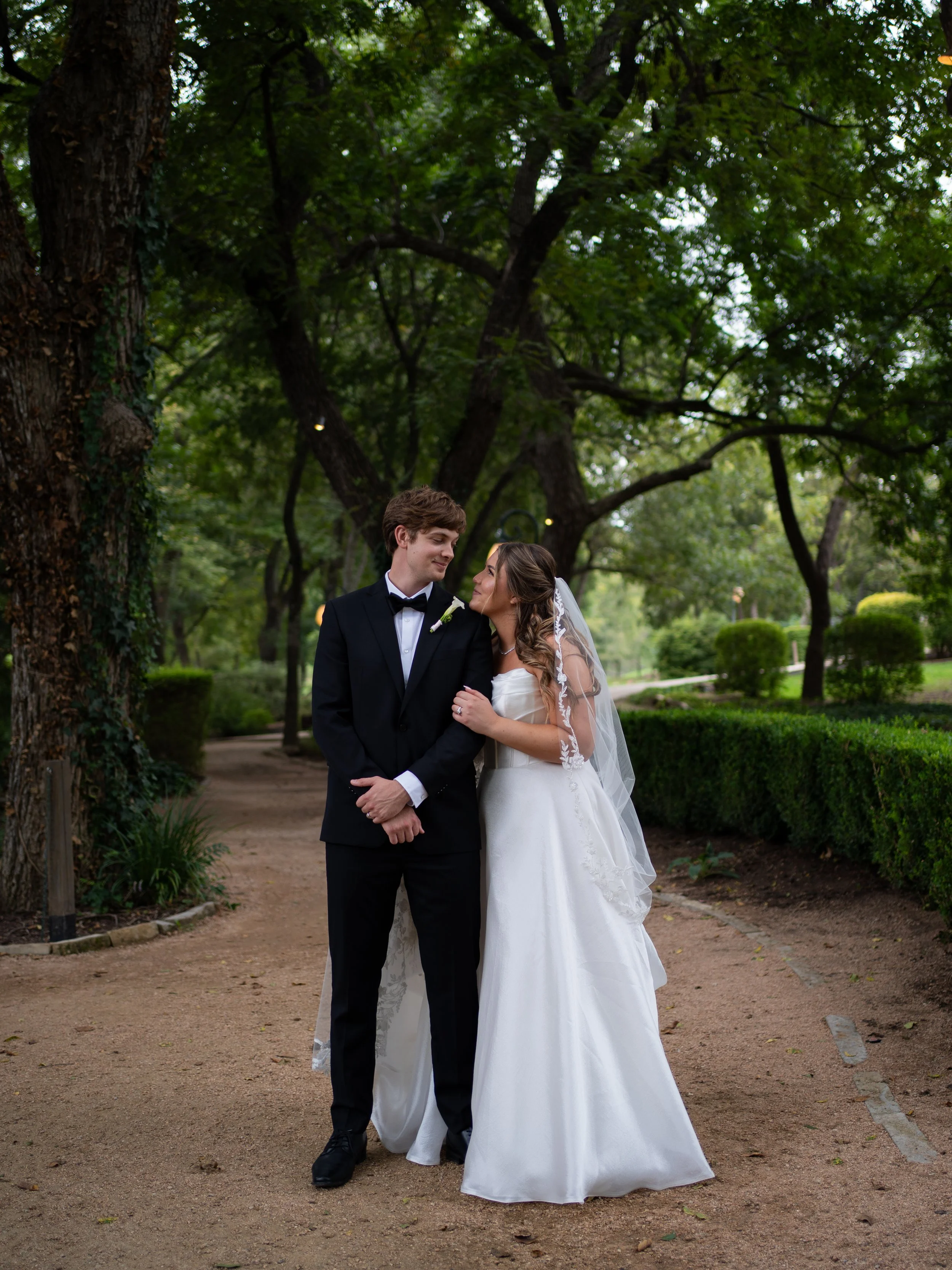 A romantic couple’s portrait in Waxahachie, Texas, framed by trees and soft natural light along a garden path.