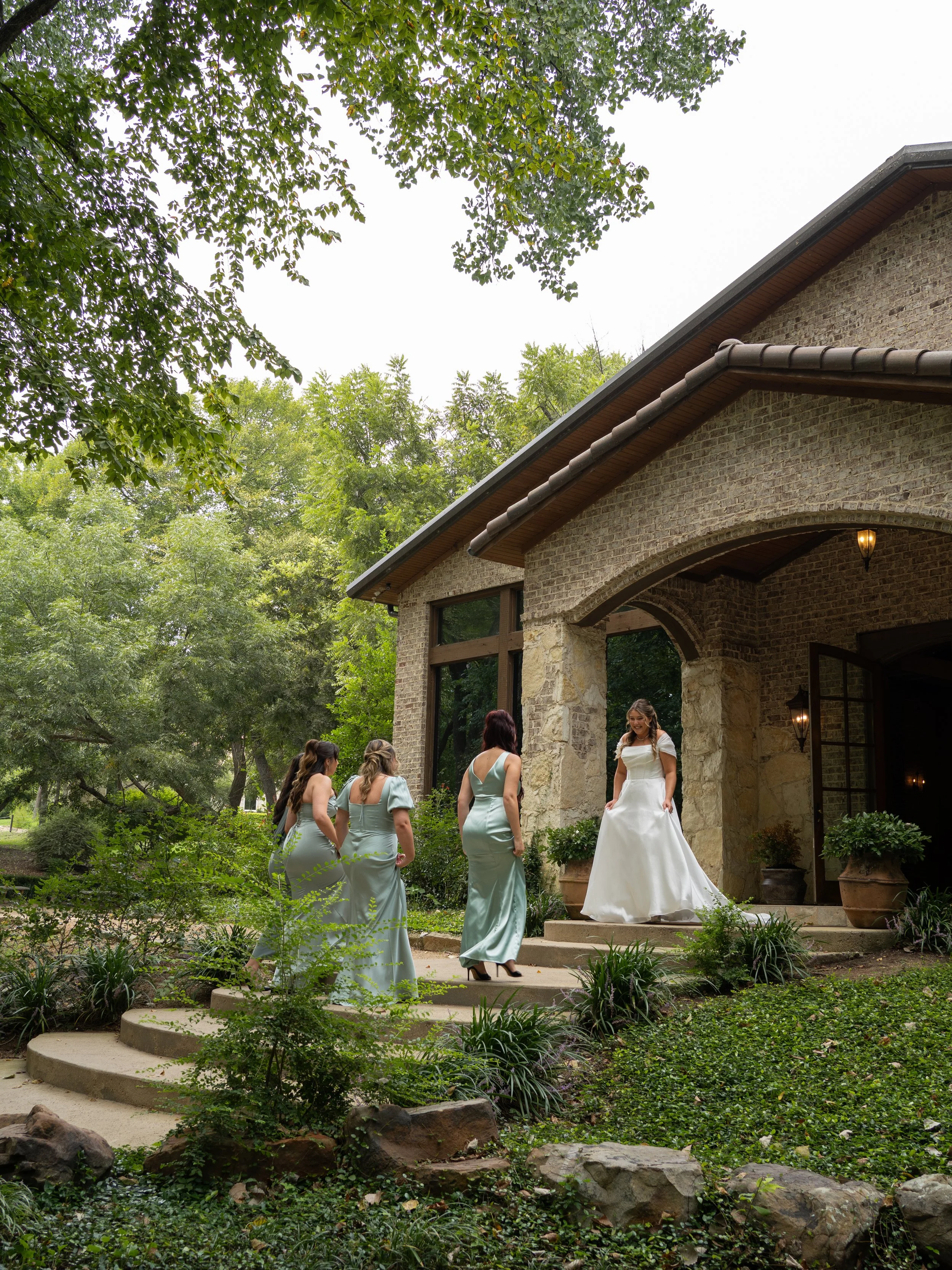 The bridal party gathers outside a Waxahachie, Texas venue, surrounded by lush greenery and natural textures.