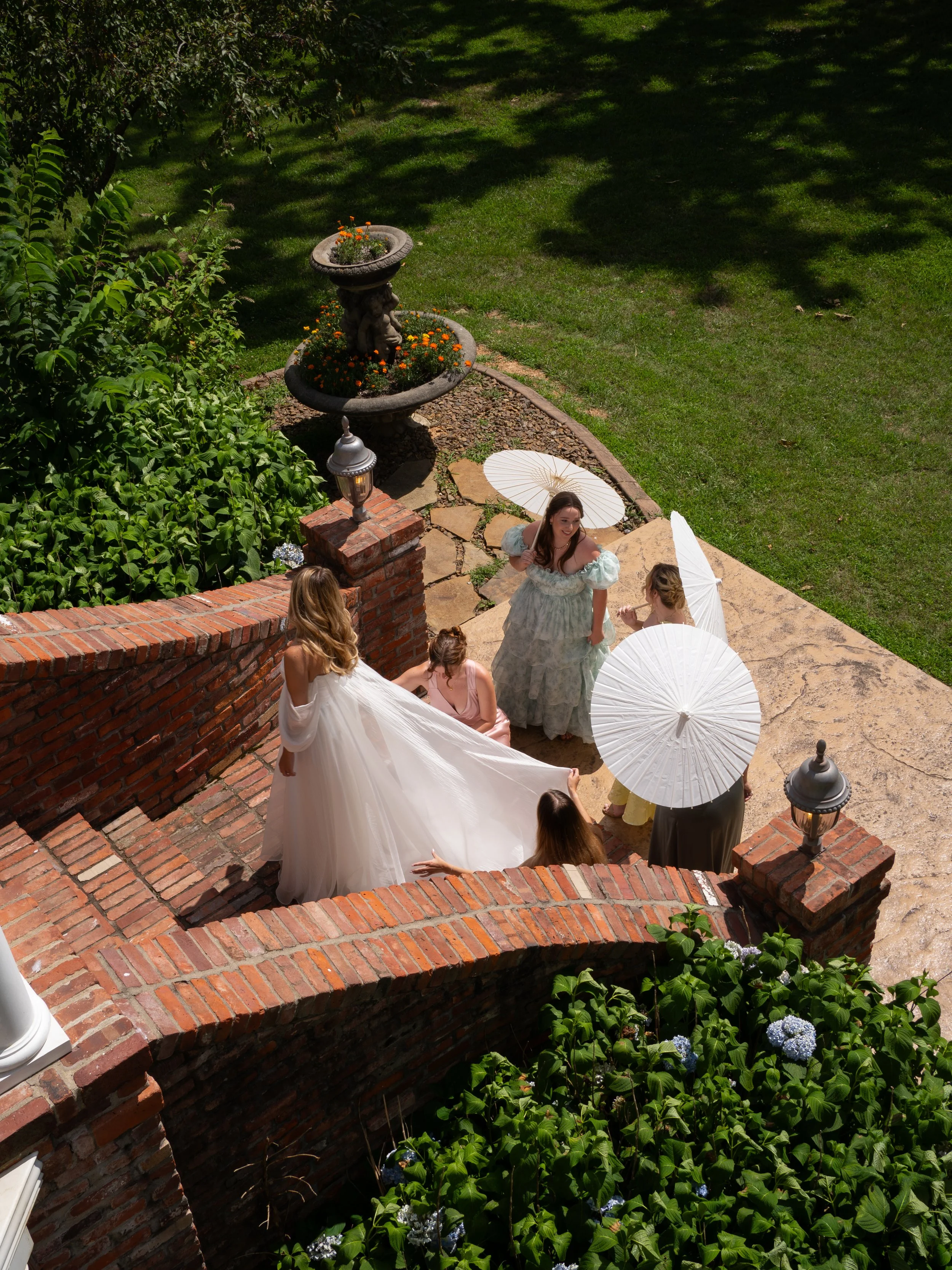 Bride and bridesmaids walking along a garden path with parasols 
