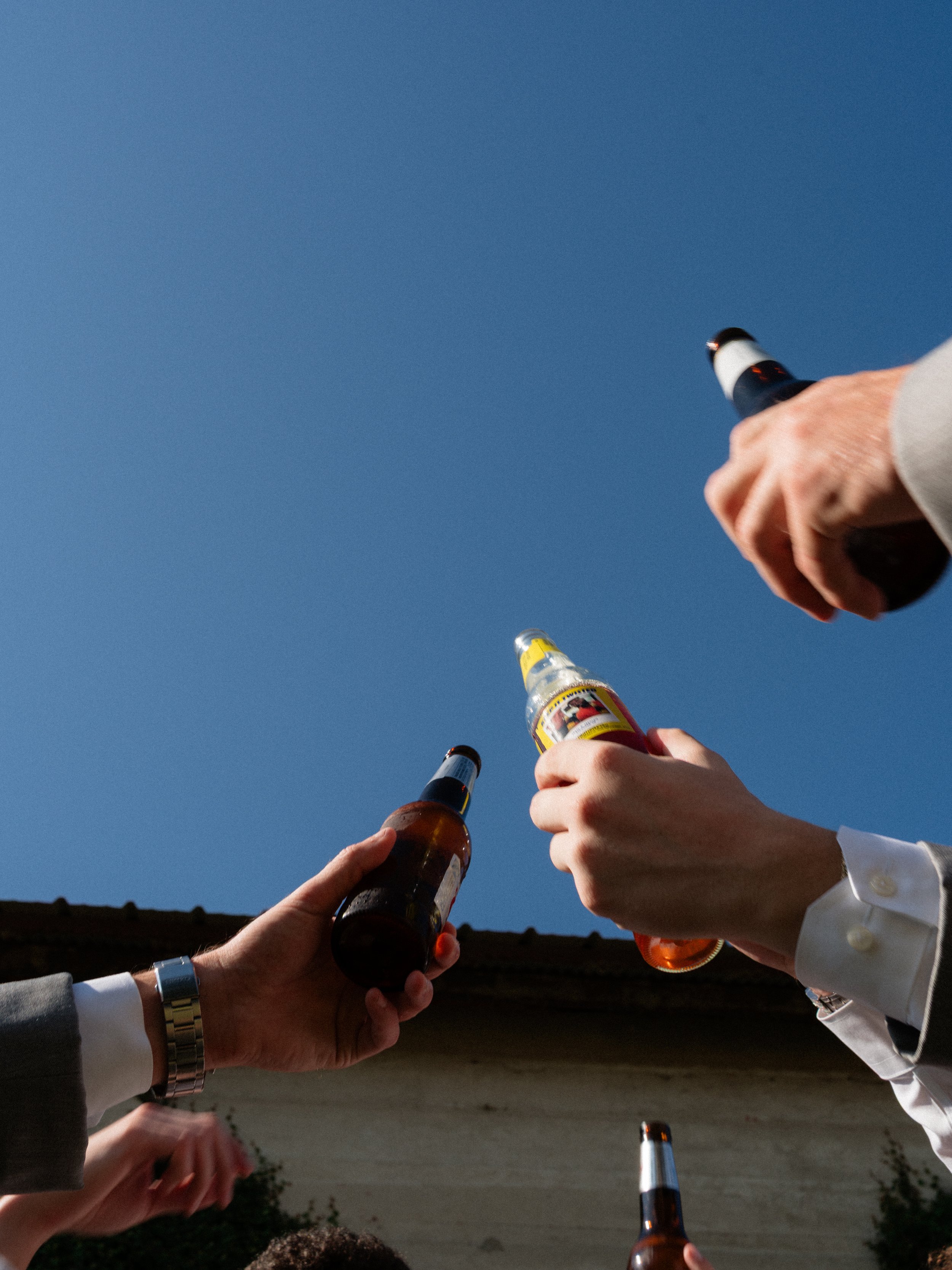 Groomsmen raising beer bottles in a celebratory toast against a blue sky at Olde Dobbin Station in Montgomery Texas
