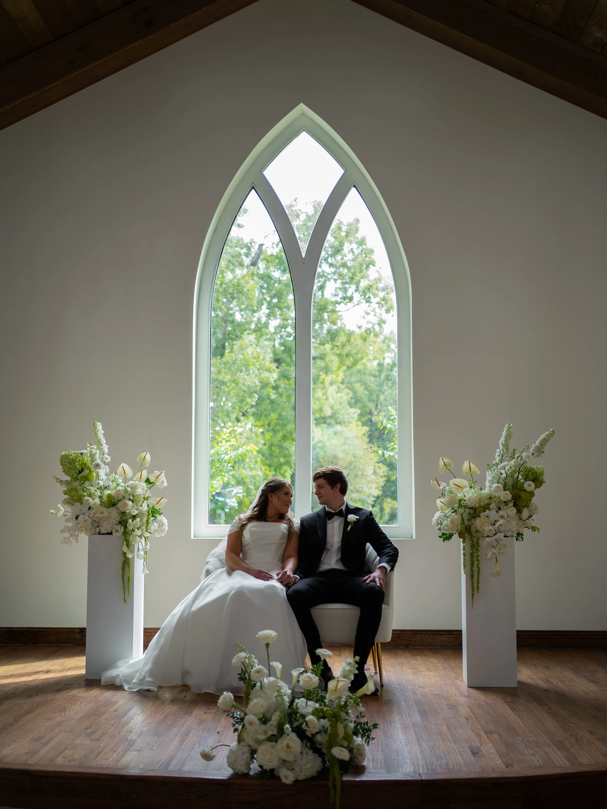 A soft and romantic portrait during wedding ceremony in Waxahachie, Texas, with the couple seated beneath an arched window framed by florals.