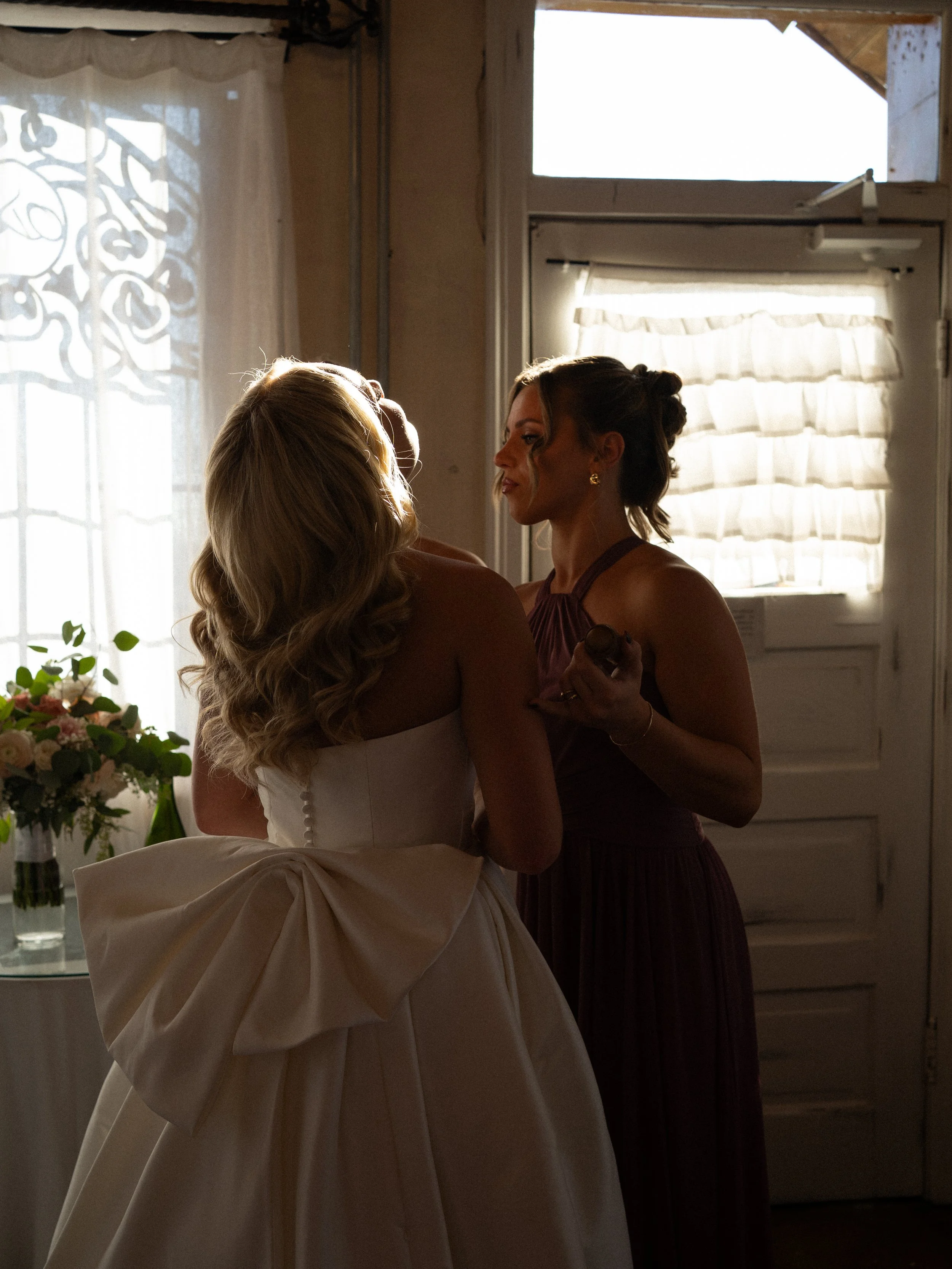 Bride getting ready with a bridesmaid in soft window light at Olde Dobbin Station in Montgomery Texas during a wedding morning