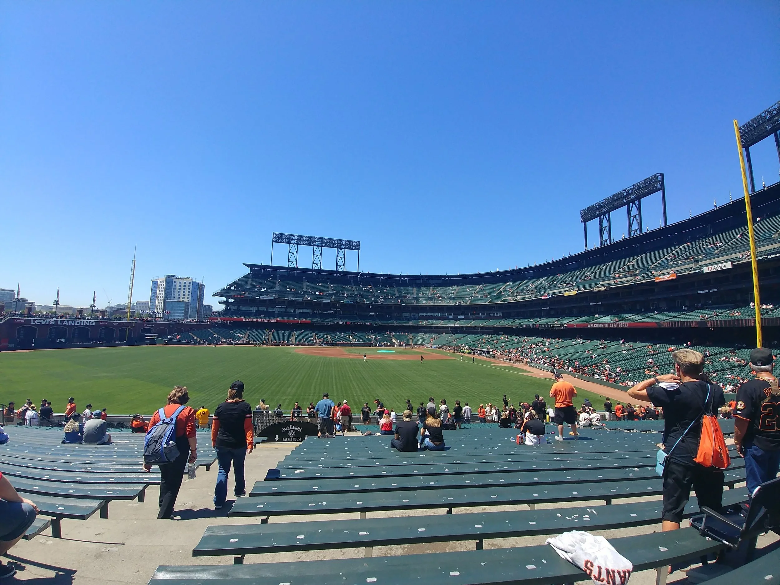 View of a baseball stadium with spectators seated in the stands on a sunny day. The field and seating area are visible, with some fans walking and some sitting. The sky is clear and blue.