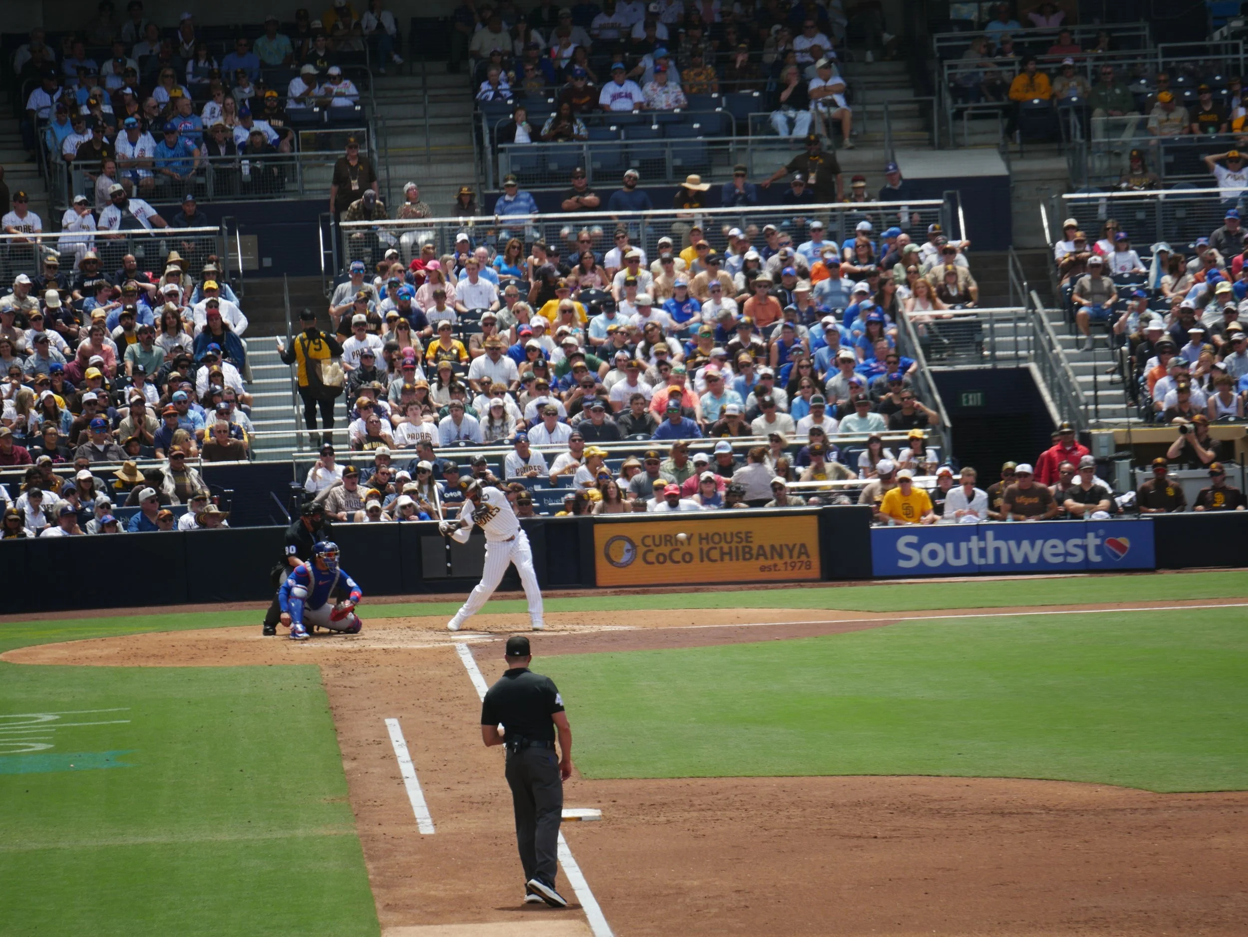 Baseball game with players at bat, pitcher on the mound, umpire behind home plate, and spectators in the stands.