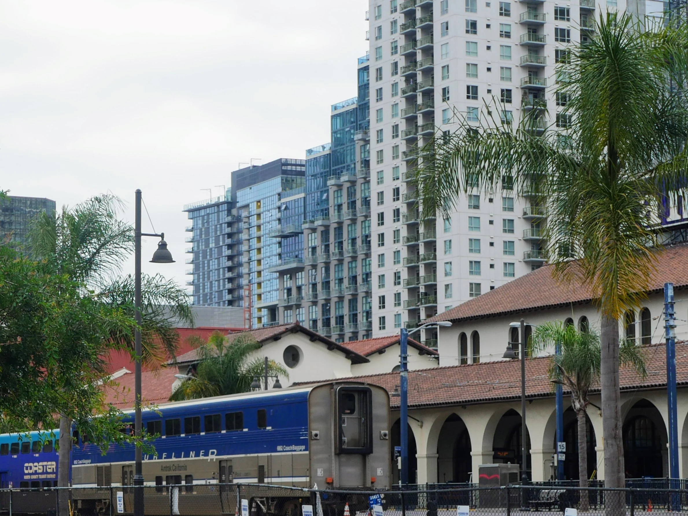 A city street scene with a double-decker bus, power lines, and tall modern apartment buildings with balconies in the background. There are trees and a red-tiled roof building in the foreground.