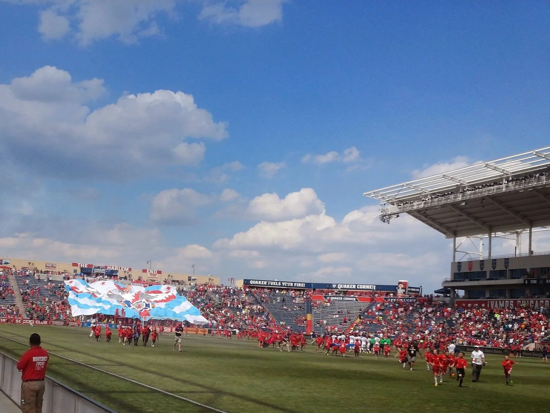 Crowd at a sports stadium with players on the field, a large American flag display, and blue sky with clouds.