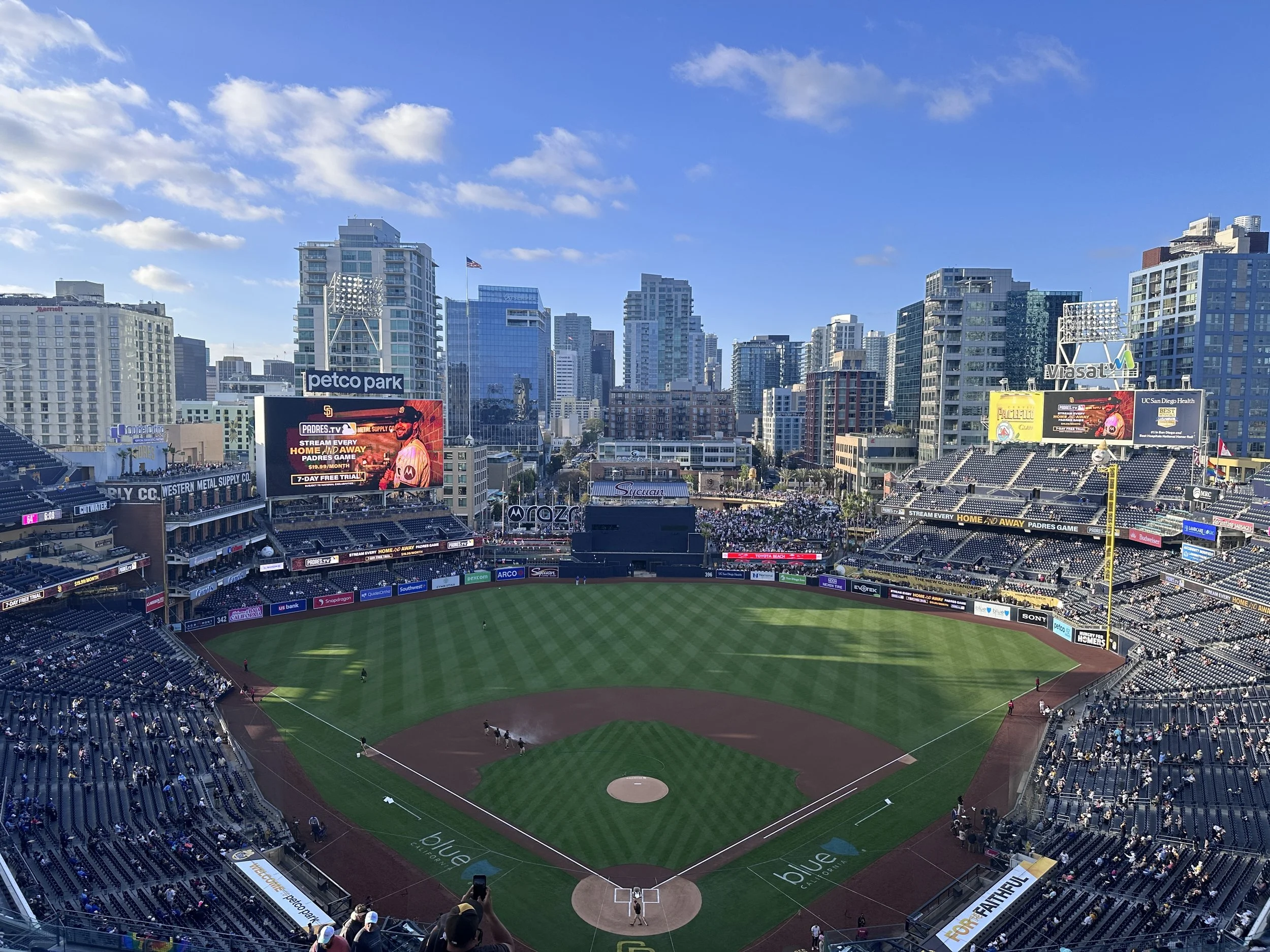 A baseball stadium filled with spectators, with high-rise buildings in the background under a partly cloudy sky.