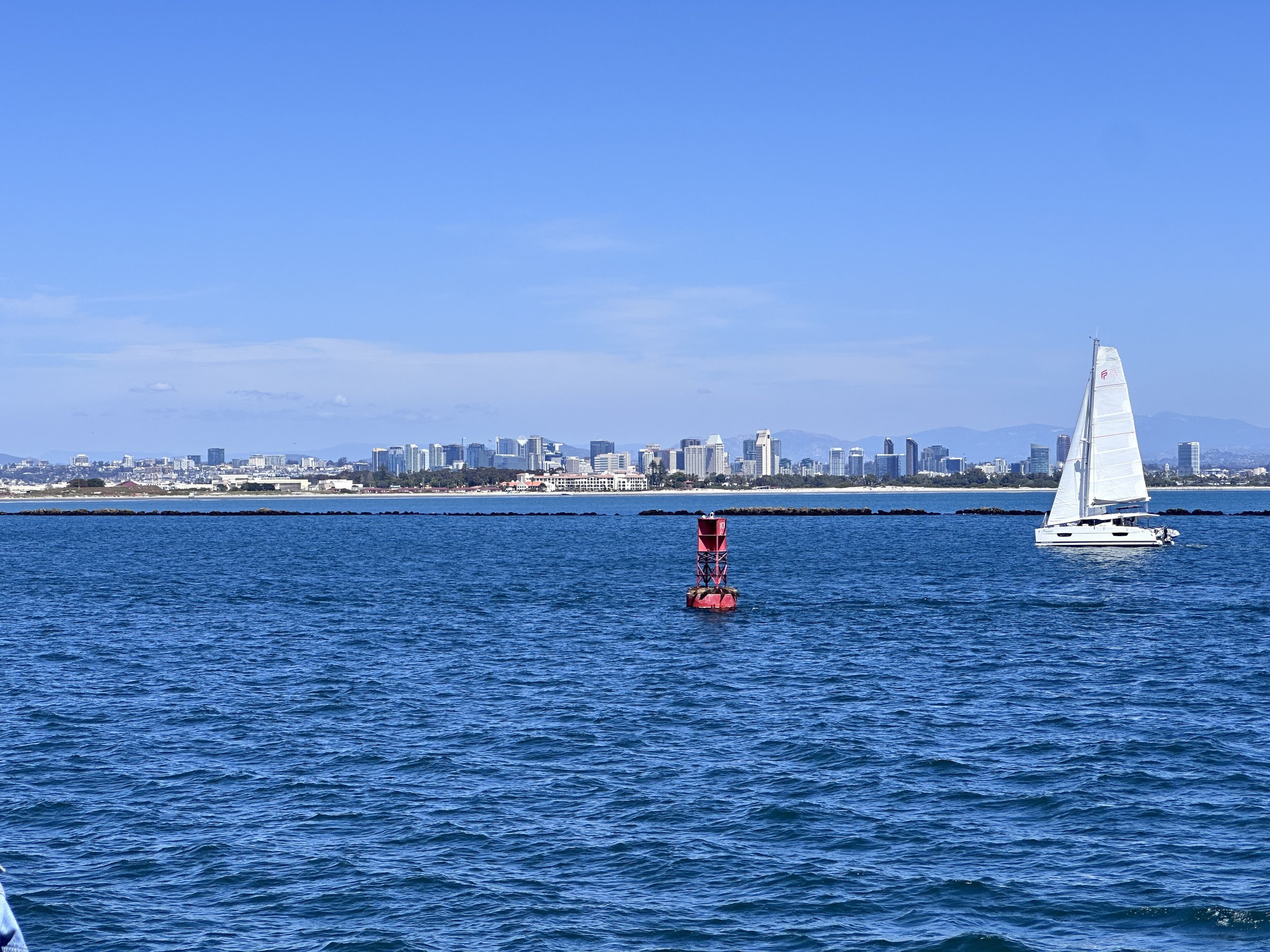 A sailboat with white sails on blue water in front of a city skyline and mountains under a clear blue sky.