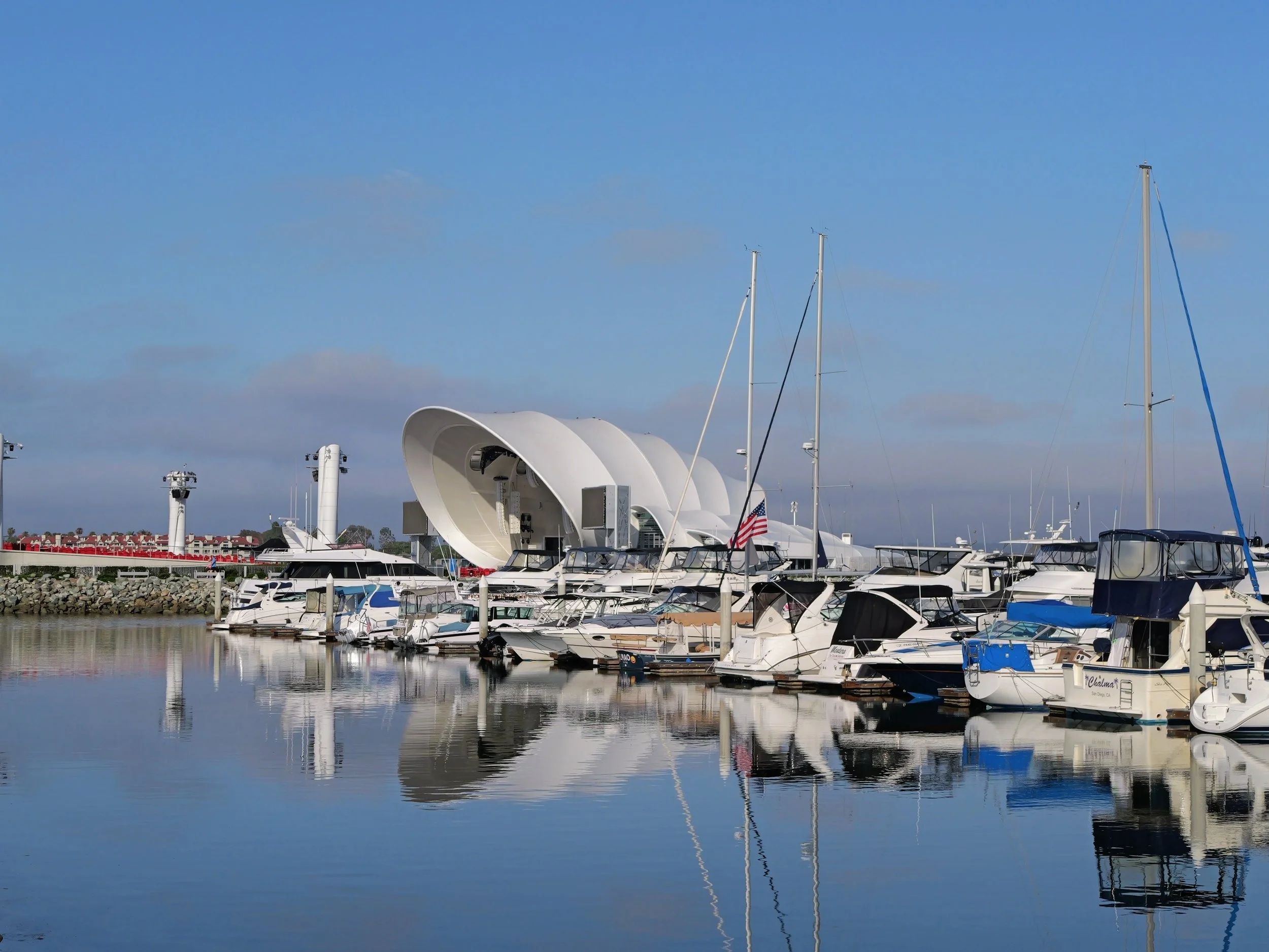 Boats and sailboats docked at a marina with calm water and a partly cloudy blue sky.