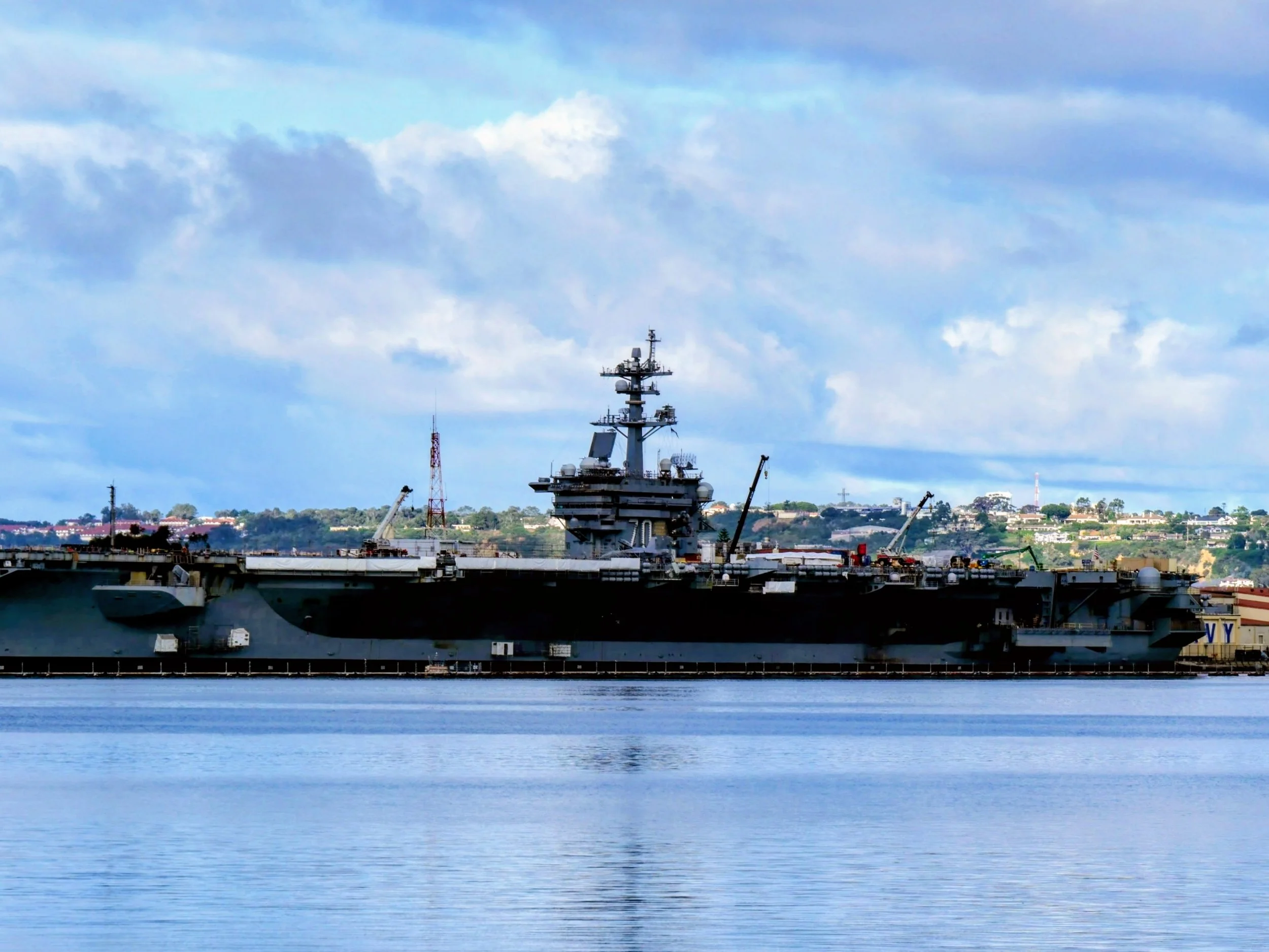A large military aircraft carrier docked in a harbor with a city and hills in the background under a partly cloudy sky.