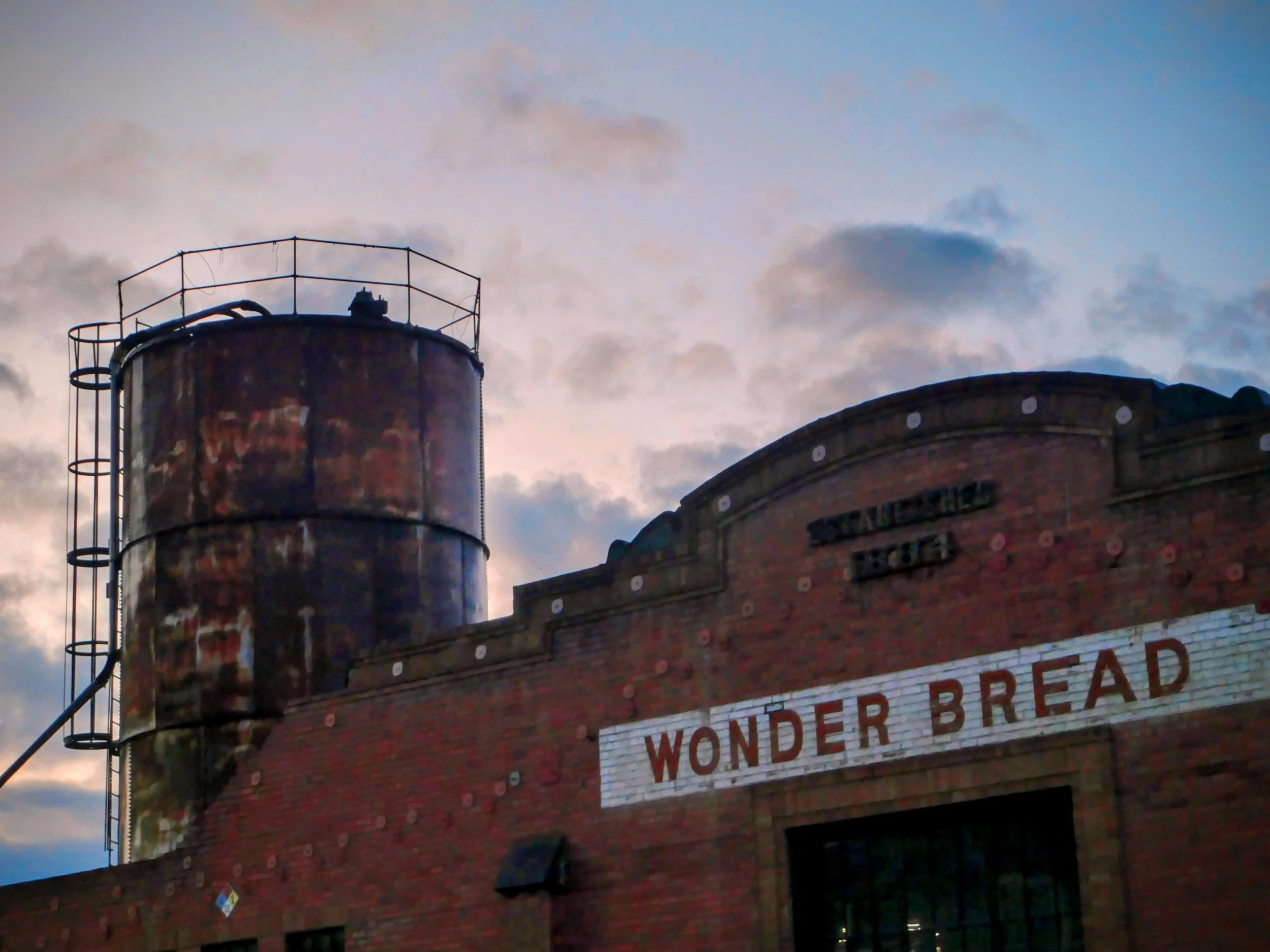 The top of a brick building with a circular tower and a rooftop railing against a sky with clouds.