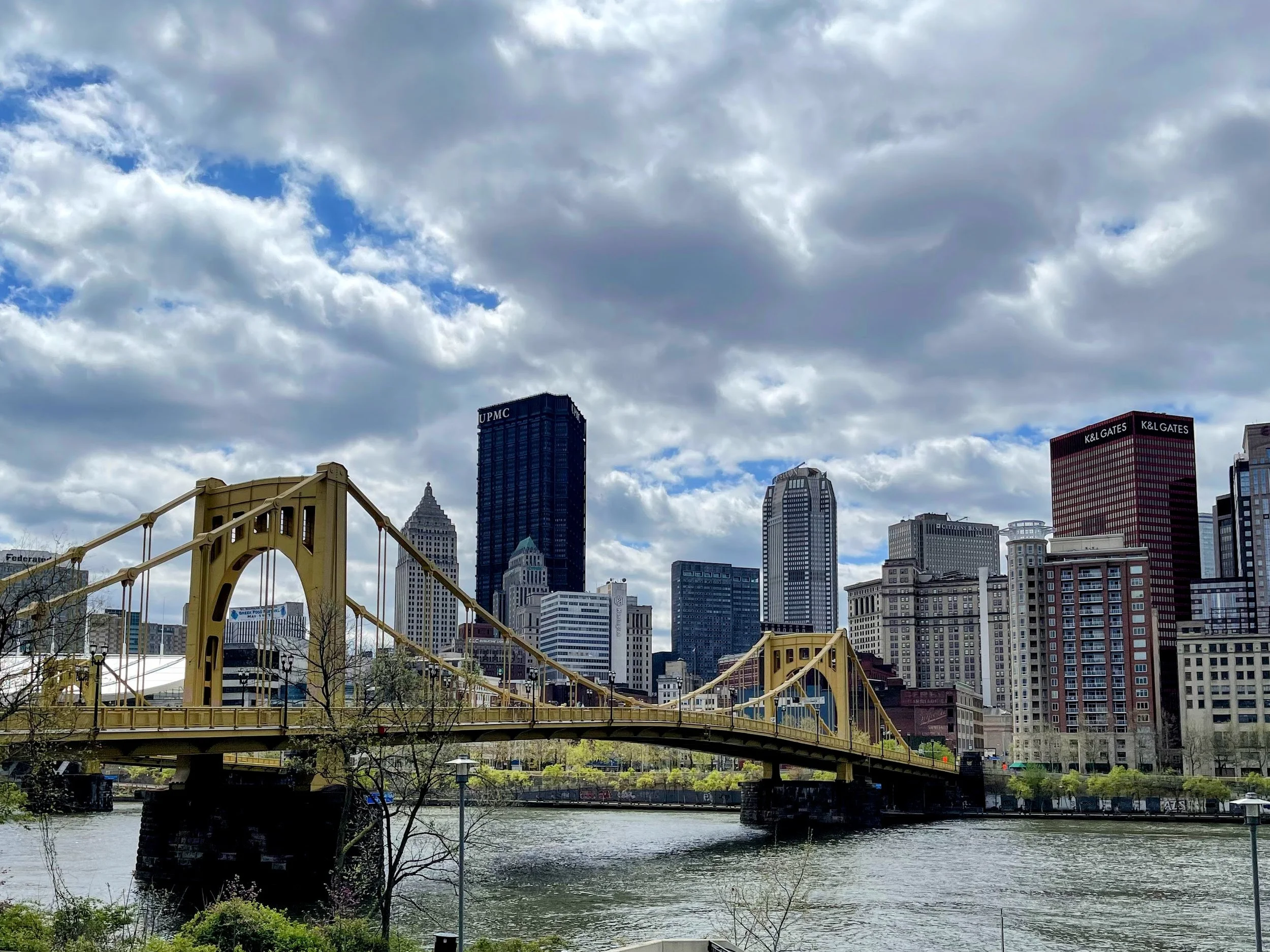 View of Pittsburgh skyline with yellow suspension bridge over river, skyscrapers under partly cloudy sky.