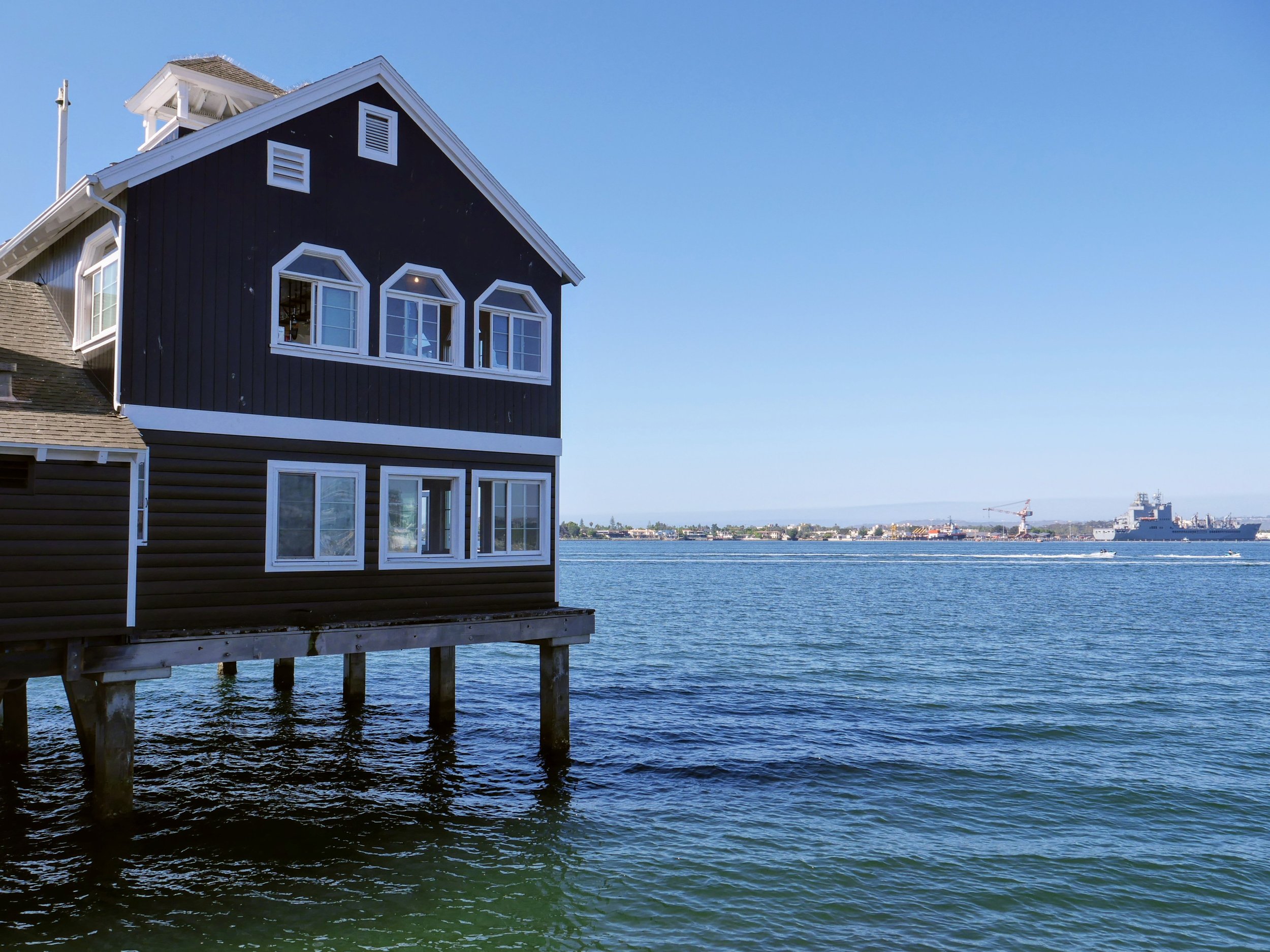 A house built on stilts over a body of water, with a ship and a crane in the distance under a clear blue sky.