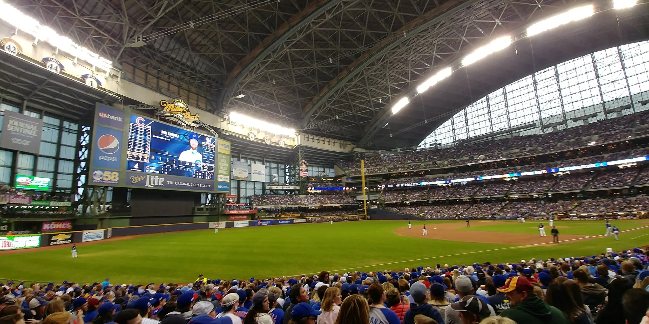 An indoor baseball stadium filled with spectators, with players on the field and a large video scoreboard showing a player's profile.