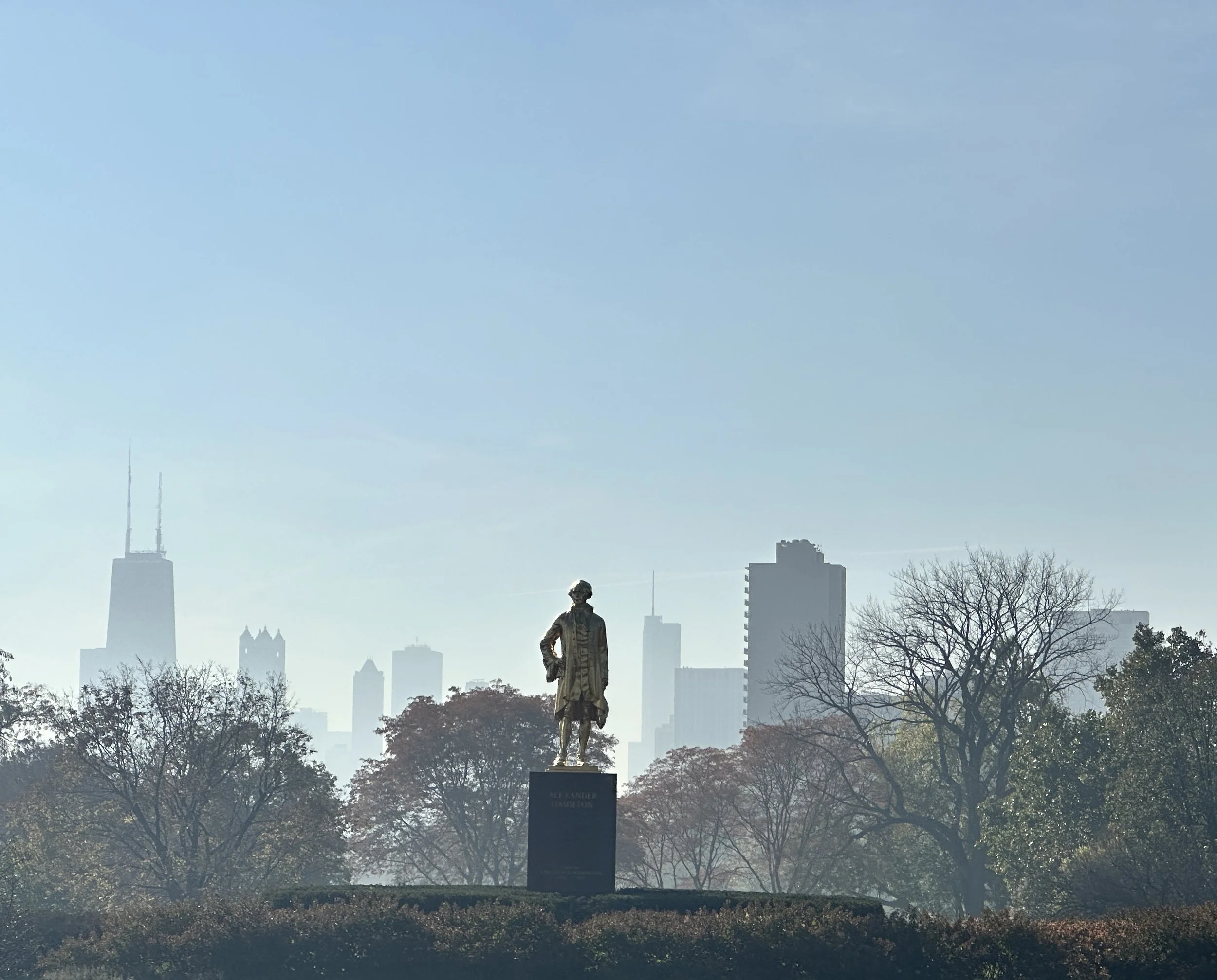 Statue of Alexander Hamilton in a park with trees and a city skyline in the background on a clear day.
