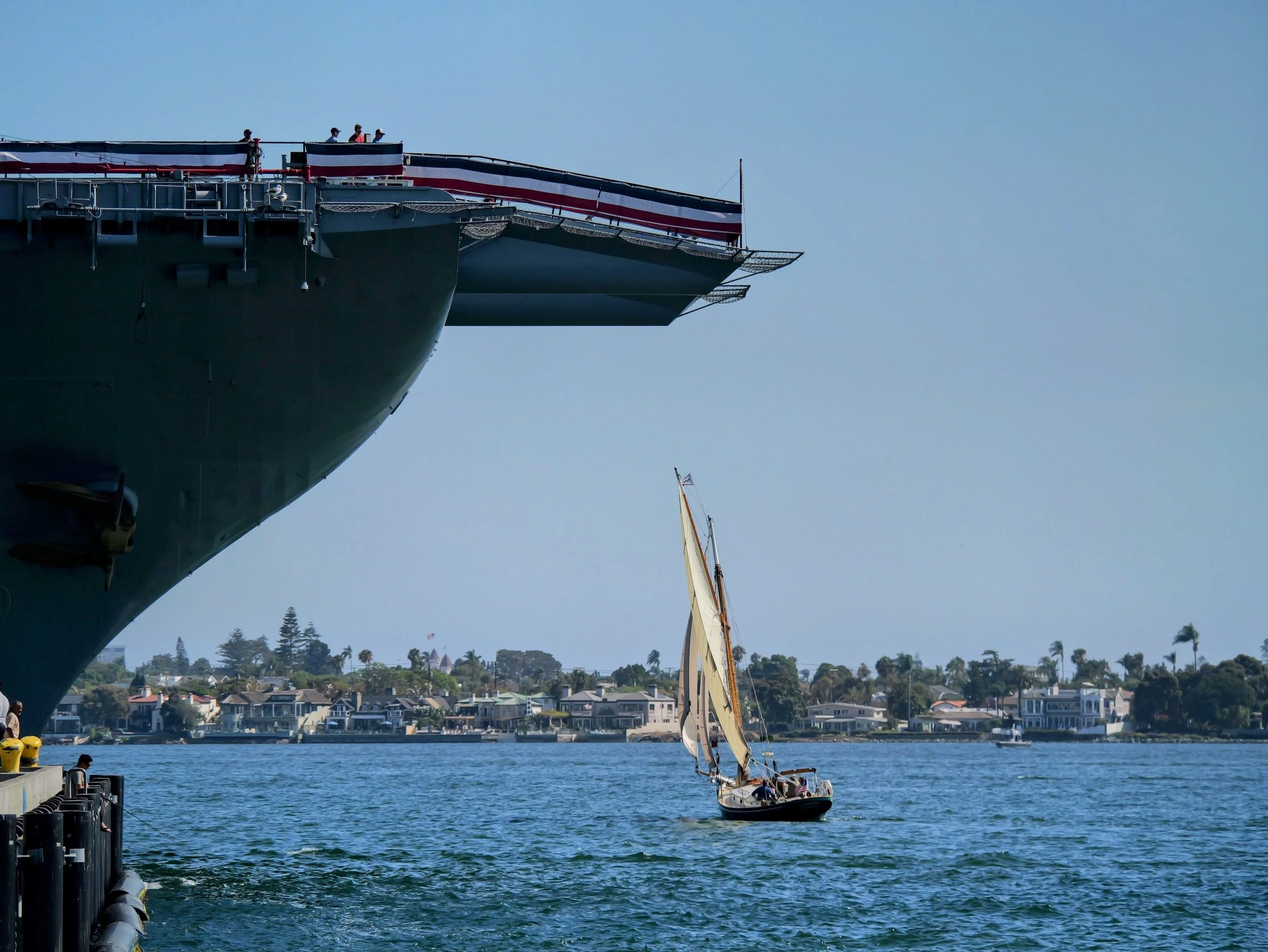 Large naval ship docked at harbor with a American flag hanging from its side; a sailboat with white sails and some houses along the shoreline in the background.